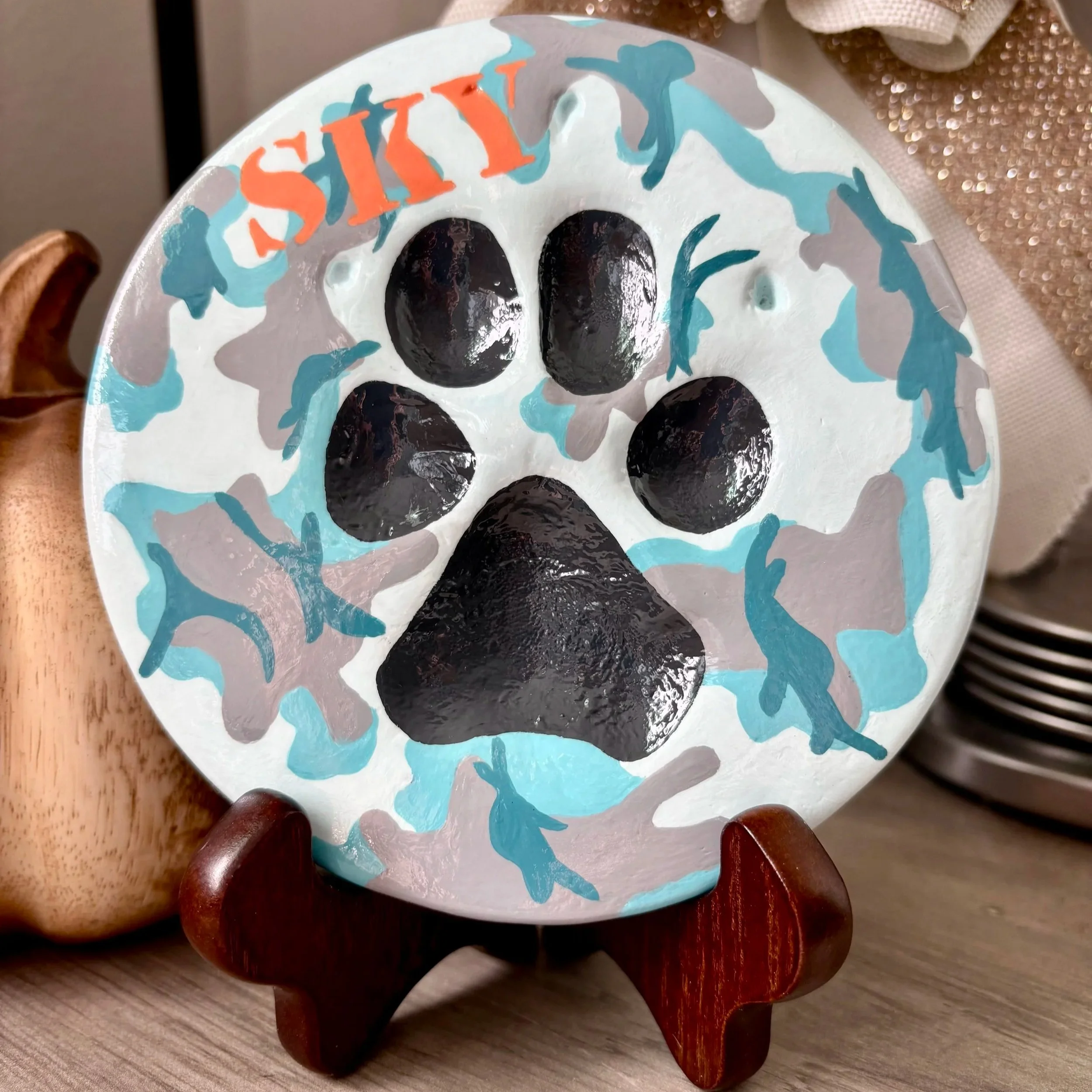 A painted paw impression where the paw pads are painted charcoal grey and the main design is a teal blue and gray camouflage, and the name 'SKY' is painted orange at the top, with holiday decor in the background.