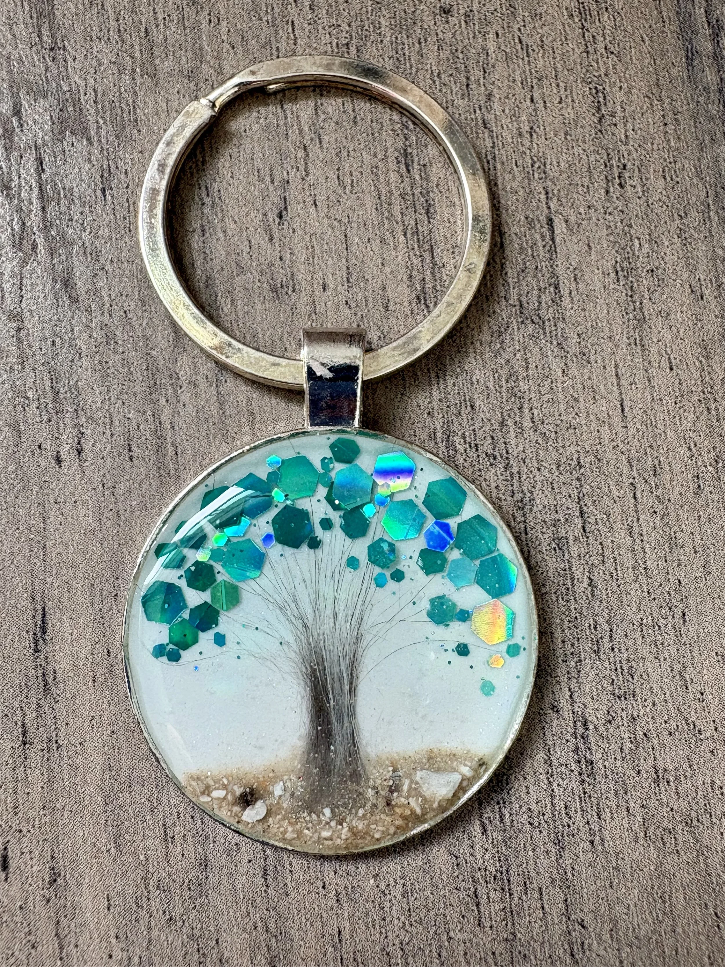 A circular keychain and ring where there is a tree-of-life encapsulated in resin to preserve pet's ash as the earth, a lock of fur for the trunk and chunky holographic chunky teal glitter for the leaves, resting on a wooden surface.