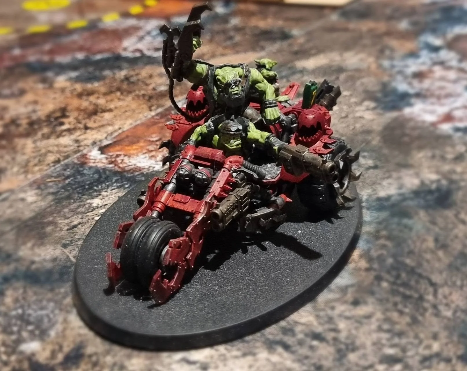 Miniature model of a green orc riding a red warthog-like vehicle with weapons, on a circular black base.