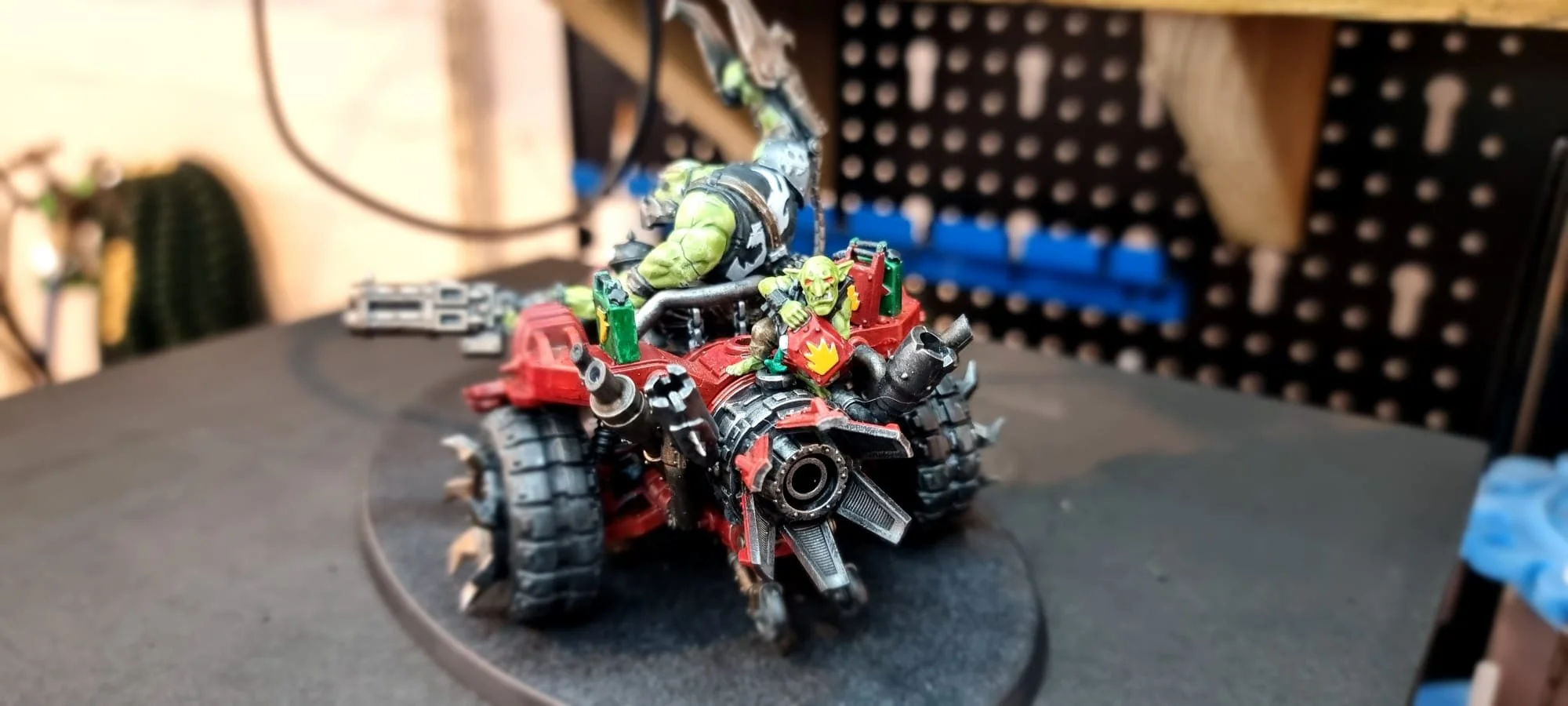 A detailed model of a war vehicle with figurines of orcs on it. The vehicle has large treaded wheels, a red and black color scheme, guns, and pipes. The orcs have green skin and are equipped with weapons. The background shows a workspace with tools and supplies.