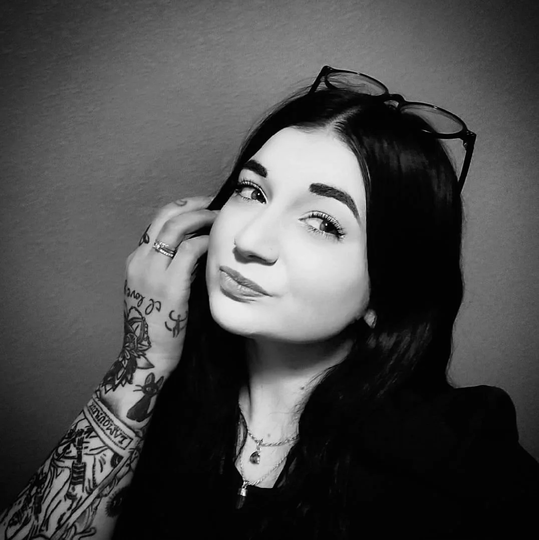 Black and white portrait of a woman with long dark hair, glasses resting on her head, and tattoos on her arm, looking at the camera with a slight smile.
