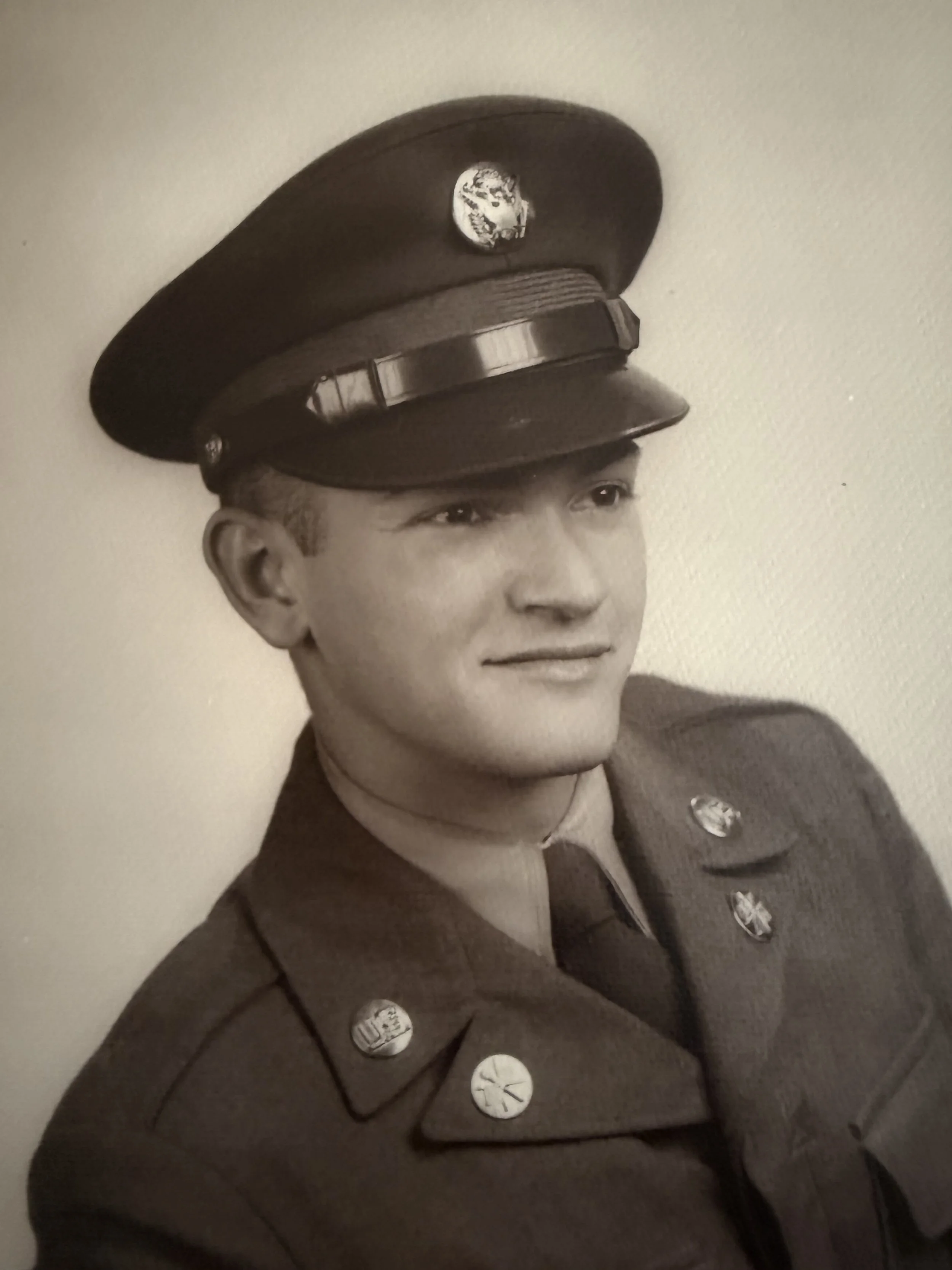 Black and white photograph of a young man in a military uniform with a cap that has an emblem, possibly a skull. The uniform has several decorative buttons and a patch on the shoulder. The young man is looking to the side with a slight smile.