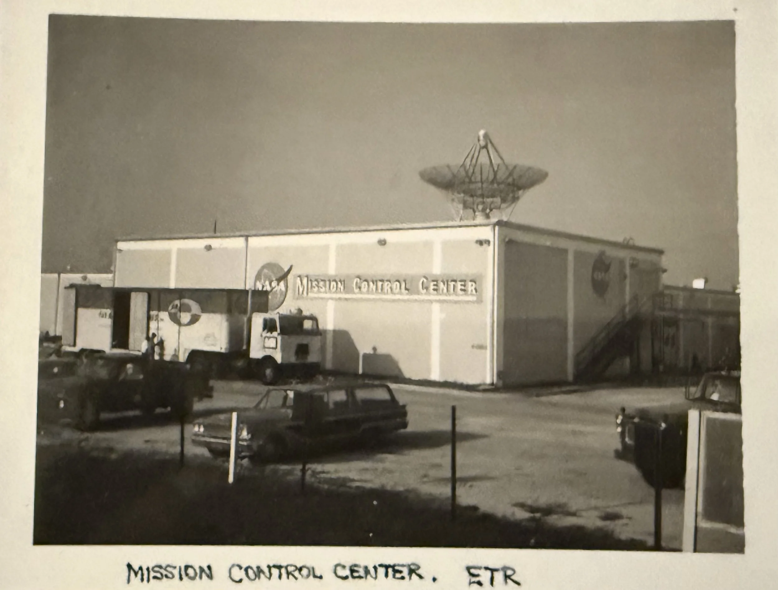 Black-and-white photo of a mission control center with a satellite dish on the roof, a van in front, and a sign reading 'Mission Control Center'.