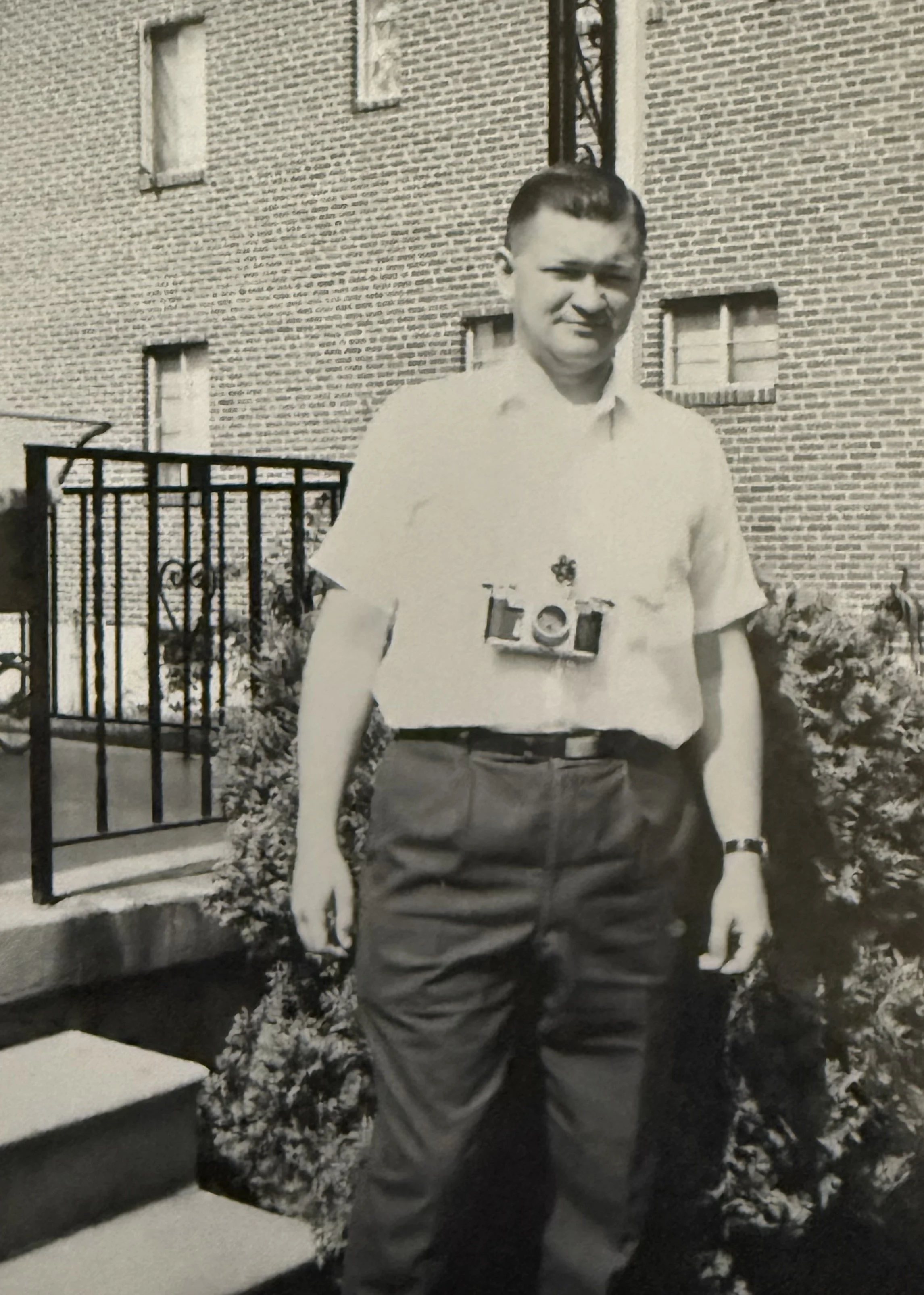A young man standing outdoors in front of a brick building with multiple small windows, wearing a collared shirt, dark pants, and a wristwatch. He has a camera hanging around his neck.