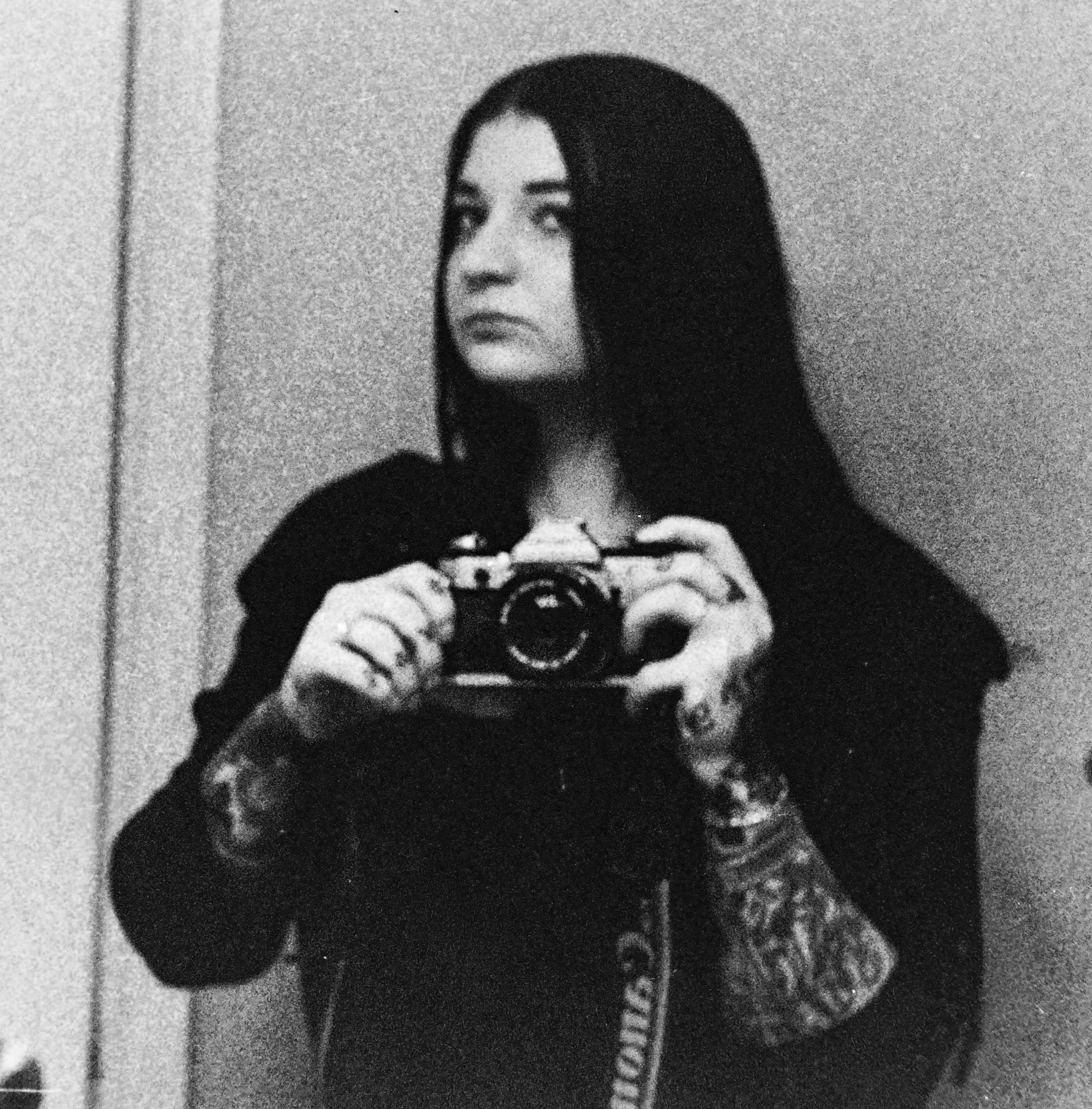 A black-and-white photograph of a woman with long dark hair taking a selfie with a camera. She is looking into the camera with a serious expression. The background appears to be a plain wall.