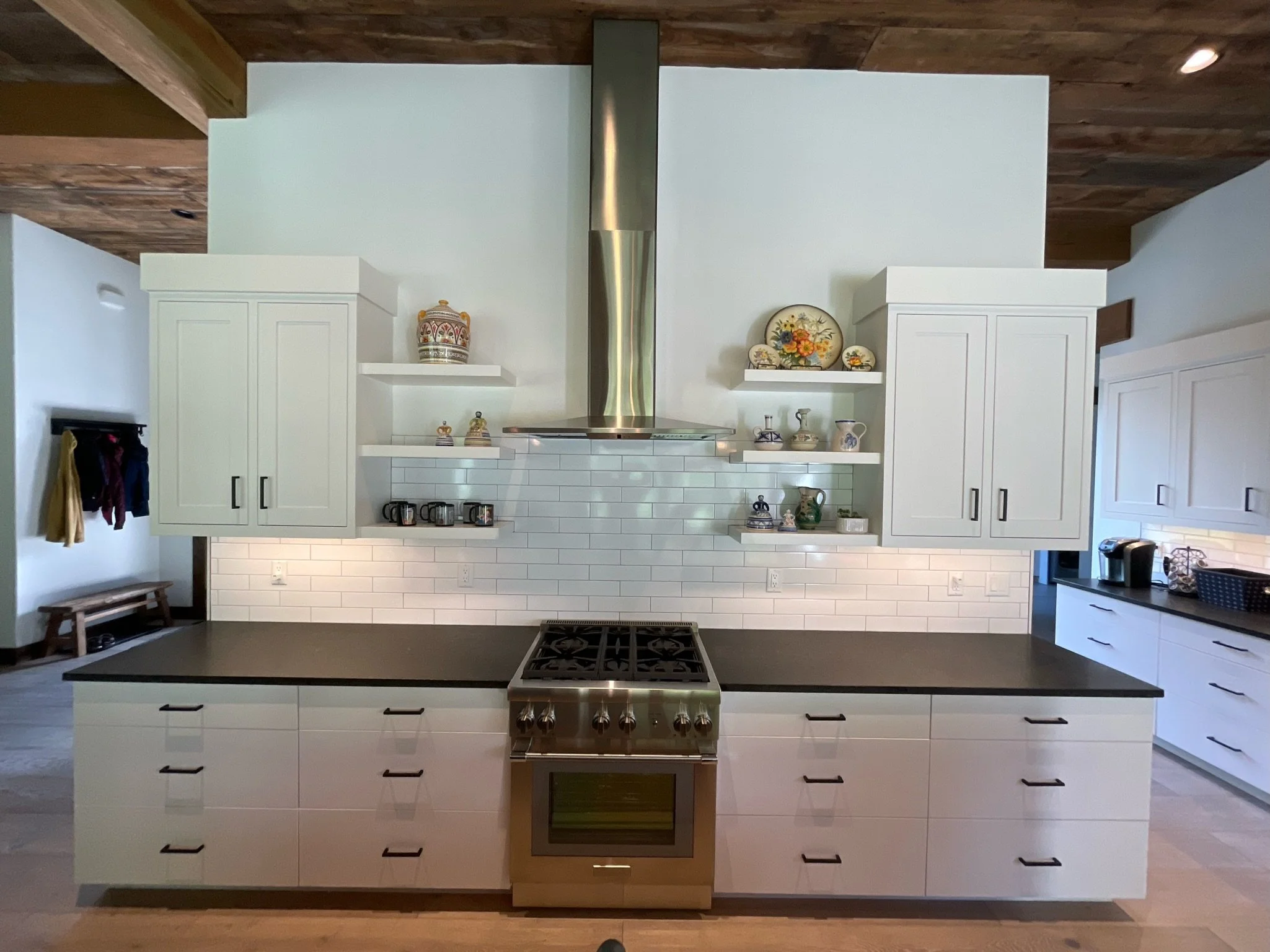 Modern kitchen with white cabinets, a black countertop, a stainless steel stove and vent, decorative plates and vases on open shelves, and a wooden ceiling.