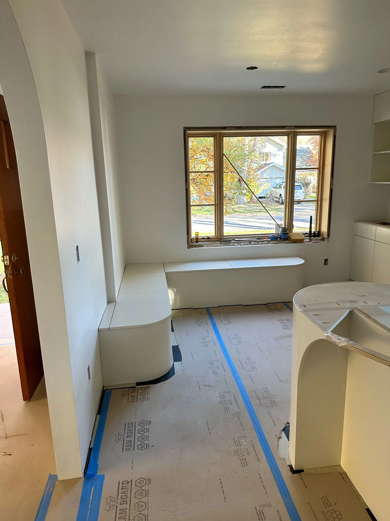 Room under construction with white cabinetry, a large window with a view of trees and houses outside, and a partially installed countertop. The floor is covered with protective paper and blue painter's tape.