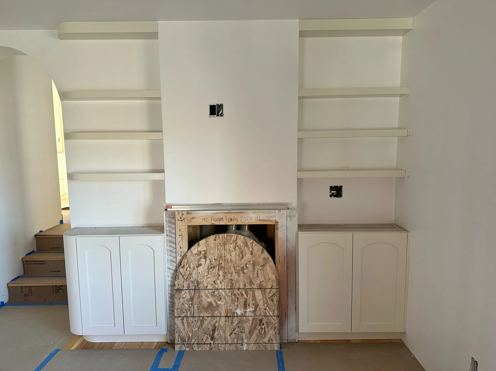 Interior wall with white cabinets, empty open shelves, and a central section under construction with plywood paneling, electrical outlets, and wiring
