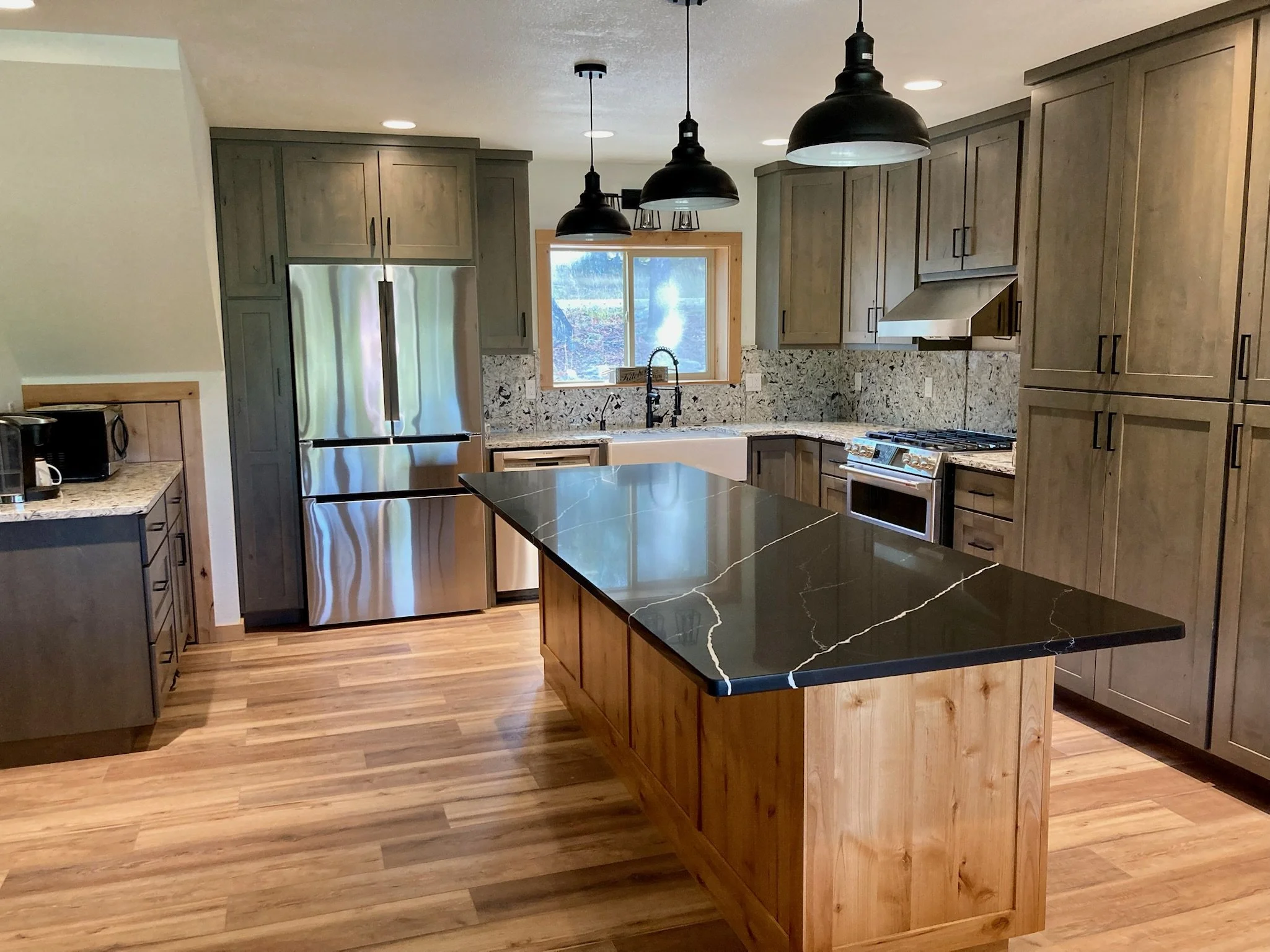 Modern kitchen with gray cabinets, black pendant lights over a black marble-island, stainless steel refrigerator, and wooden flooring, with a window above the sink.