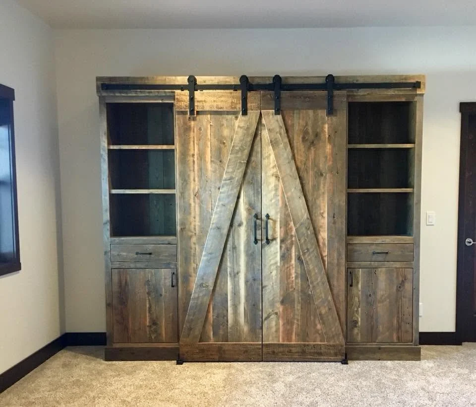Wooden barn door with sliding hardware, flanked by built-in shelves and cabinets, installed in a room with beige walls and carpeted floor.