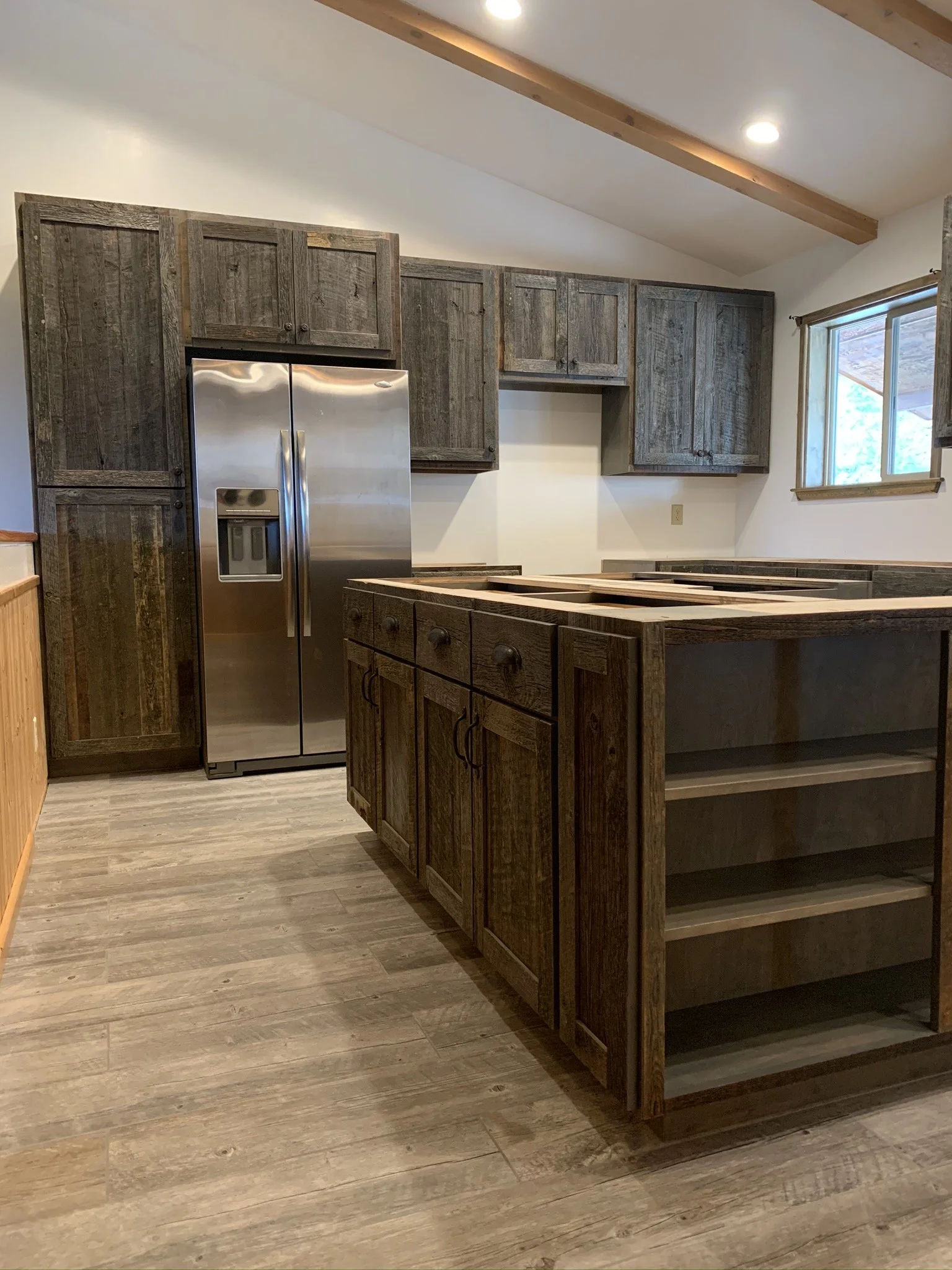 Interior view of a kitchen with wooden cabinets, a stainless steel refrigerator, and a window, under a sloped ceiling with wooden beams.
