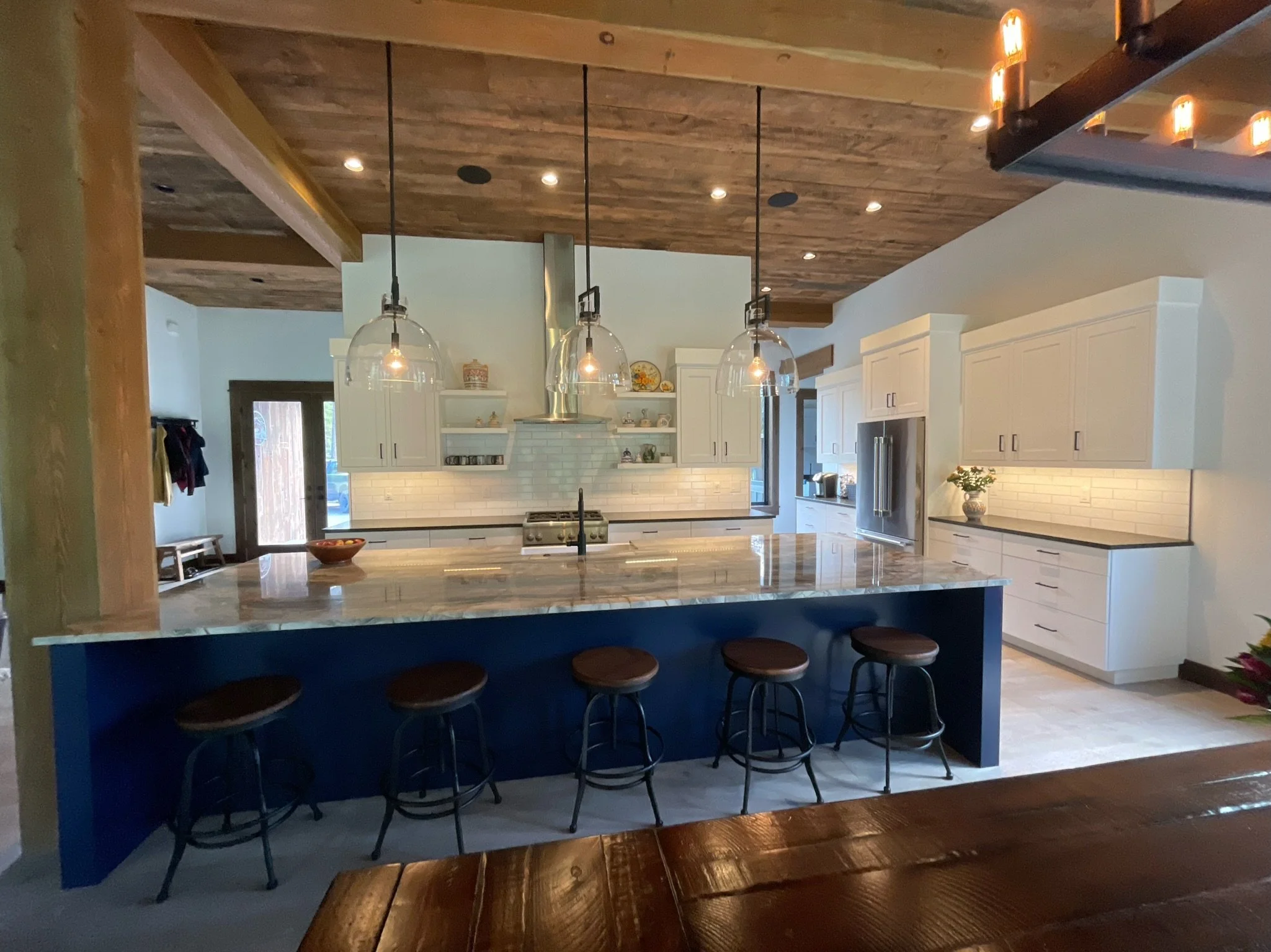 Modern kitchen with white cabinets, a marble island counter with bar stools, pendant lights hanging from a wooden ceiling, a stainless steel refrigerator, and decorative items on open shelves.