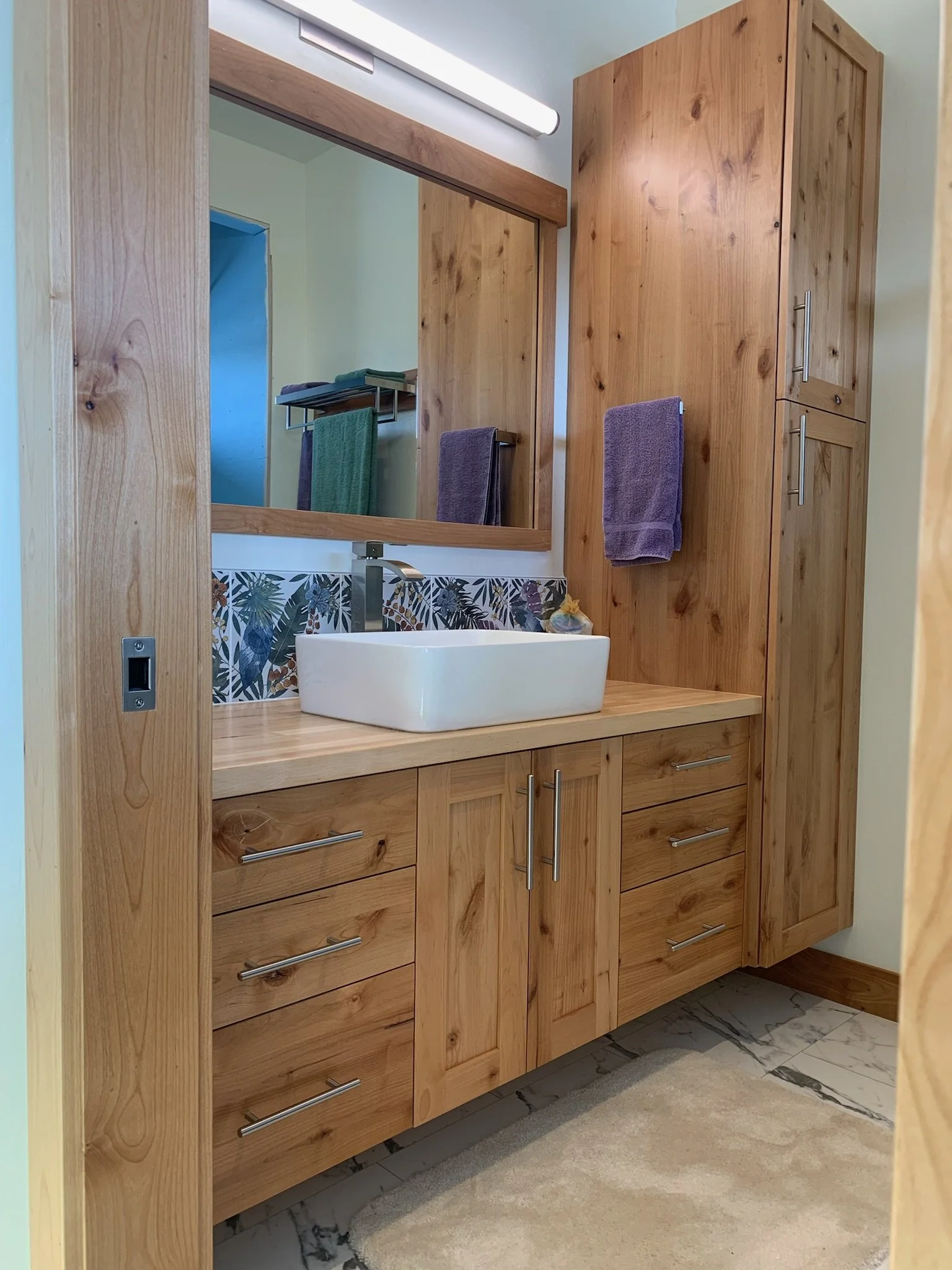 Wooden bathroom vanity with a white vessel sink, large mirror, purple towel, and a wooden cabinet to the right, with marble flooring and a light-colored wall.