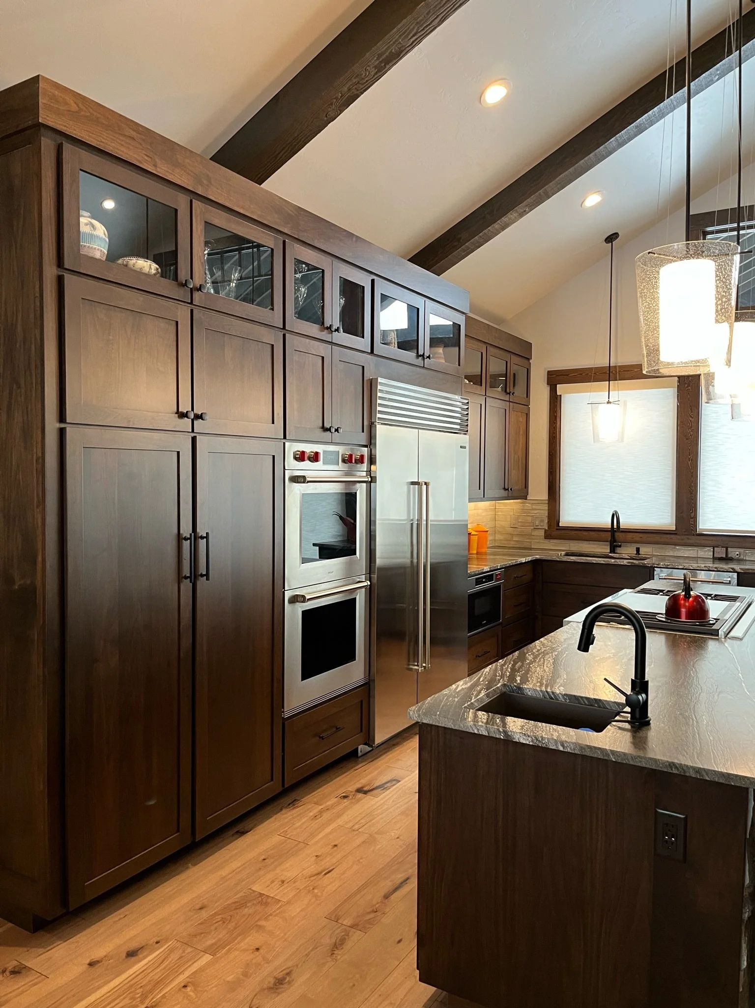 Kitchen with wooden cabinets, granite island, double oven, stainless steel refrigerator, wood floors, large window, and modern light fixtures.