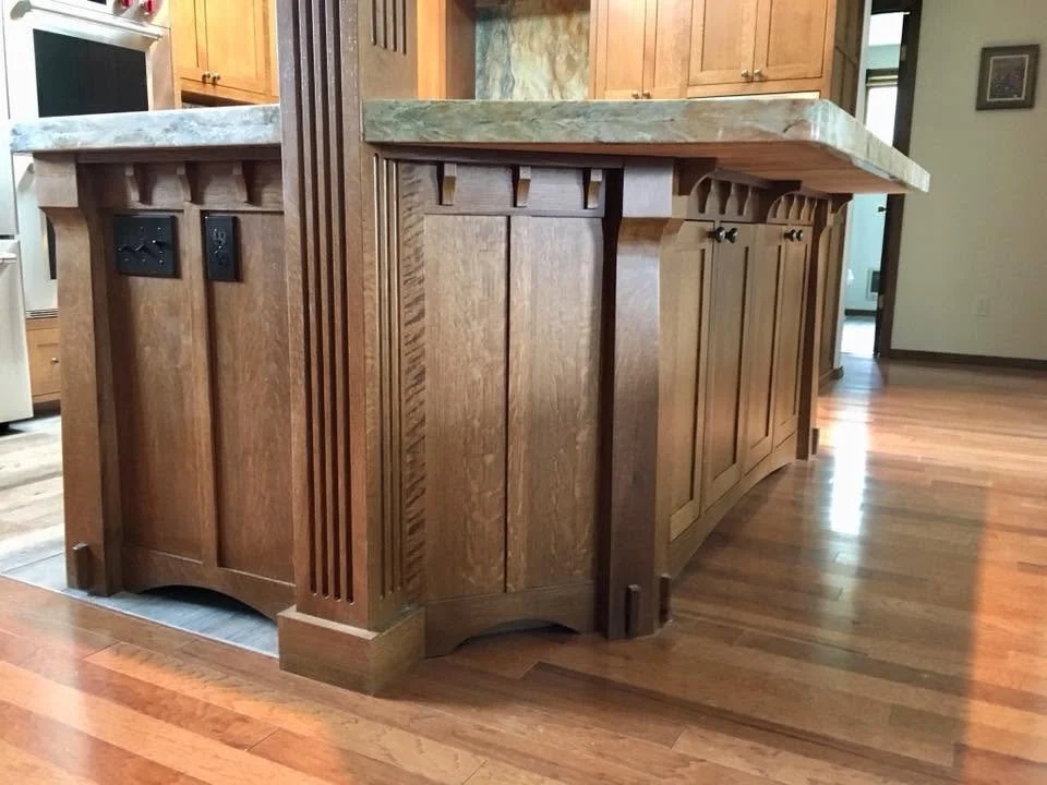 Wooden kitchen island with a light-colored marble countertop, built-in electrical outlets, and vertical slats on the sides, situated in a home kitchen with hardwood floors and wooden cabinets.