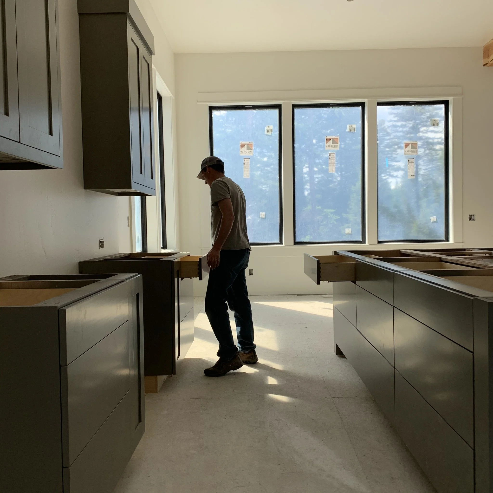 Construction worker standing in a partially built kitchen with cabinets and large windows in the background, sunlight streaming through.