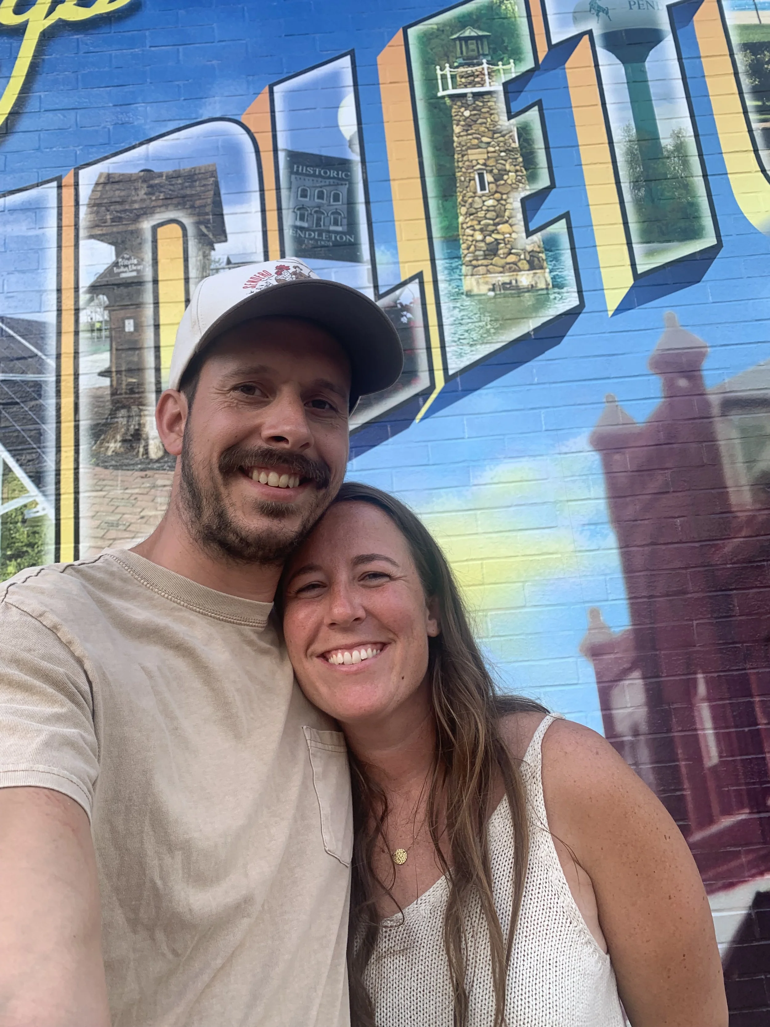 A smiling man and woman taking a selfie in front of a colorful mural with images of a lighthouse, a clock tower, and a water tower.