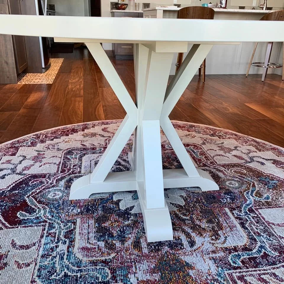 A white wooden table with a cross-legged base, placed on a patterned rug in a kitchen setting.