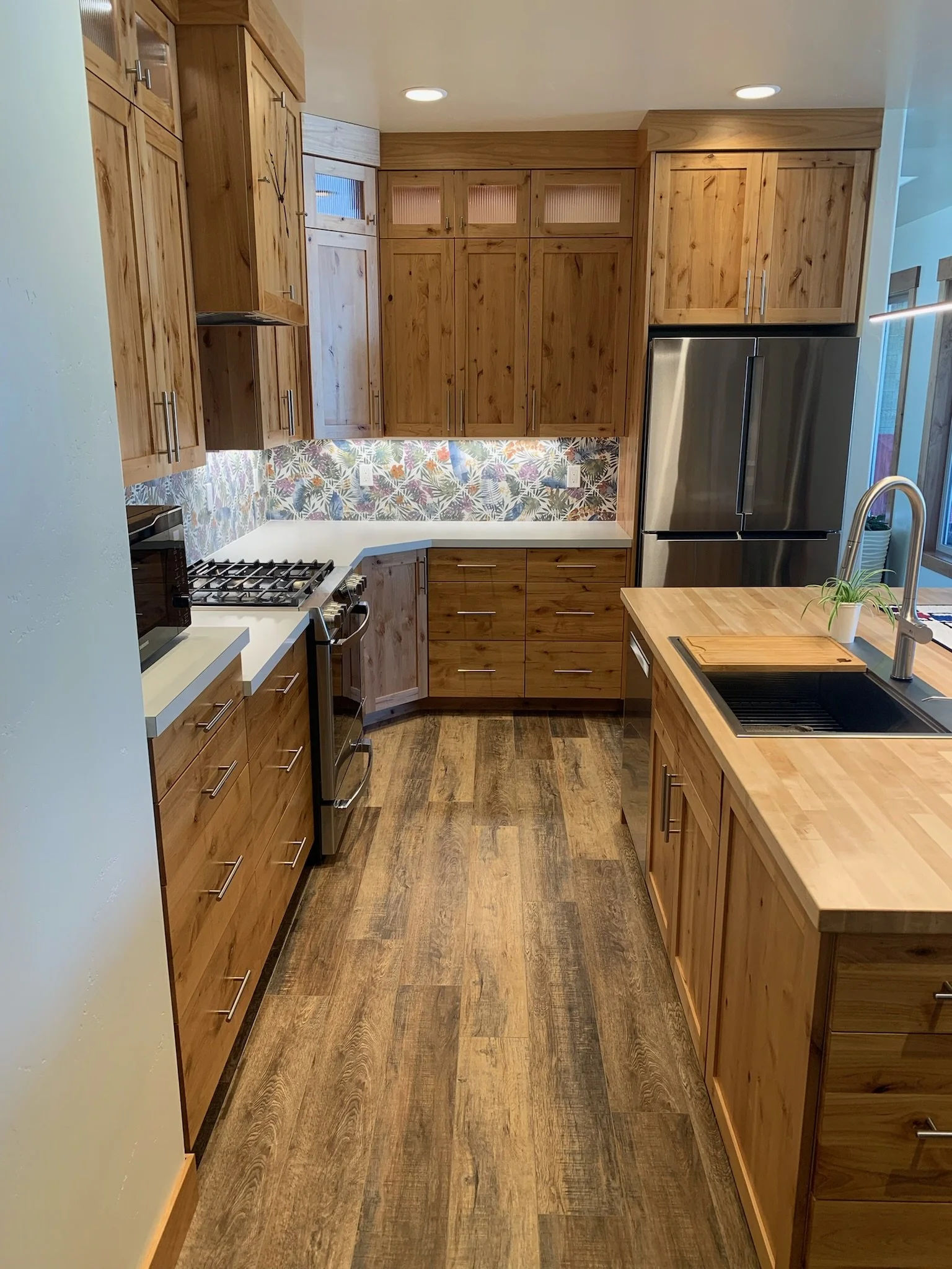 A kitchen with wooden cabinets, a stainless steel refrigerator, colorful backsplash, wooden floor, and a small plant near the sink.