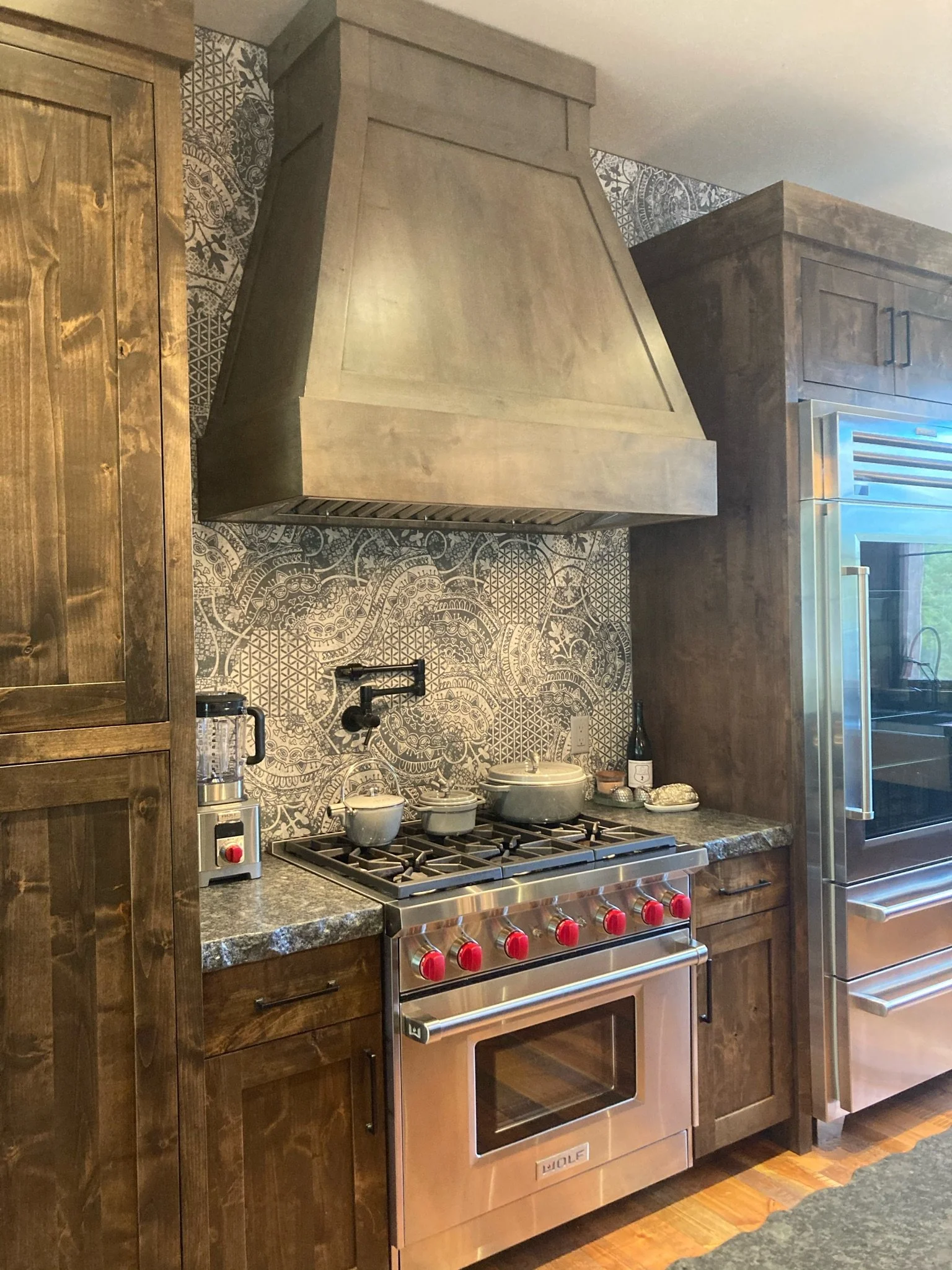 A kitchen with a stainless steel Wolf range stove, wooden cabinets, patterned tile backsplash, and granite countertop. Items on the counter include bowls, a bottle of wine, and various utensils.