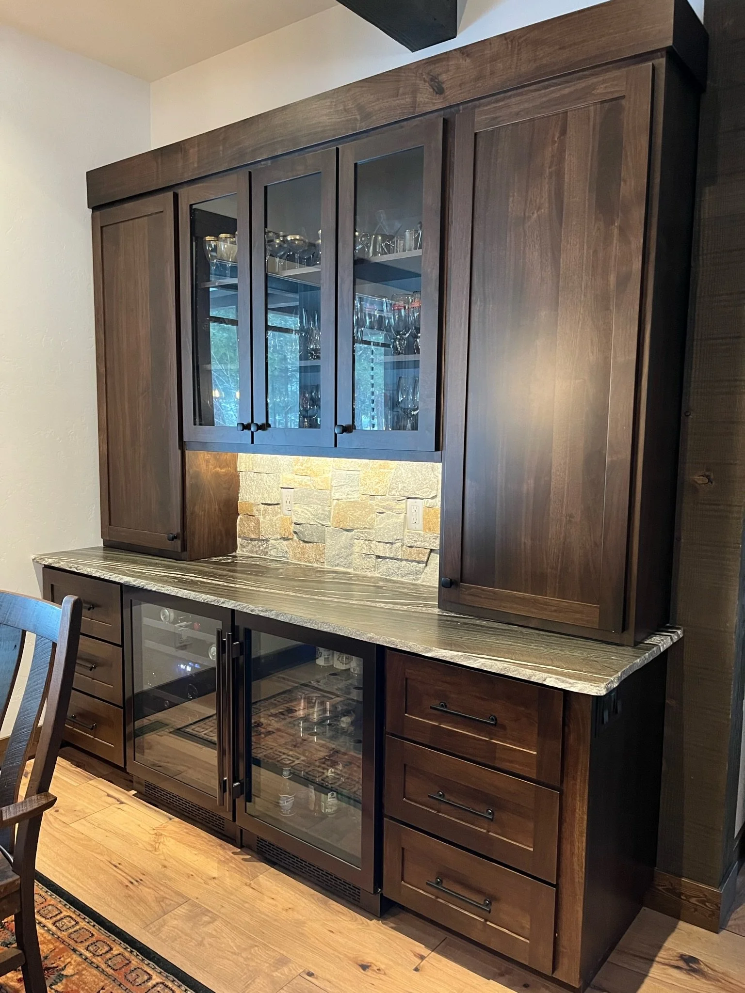 A kitchen cabinet display with glass doors showcasing glassware, including wine glasses, on a shelf. The cabinet is made of dark wood with a stone countertop beneath it. The background features a textured stone wall with electrical outlets. Part of a