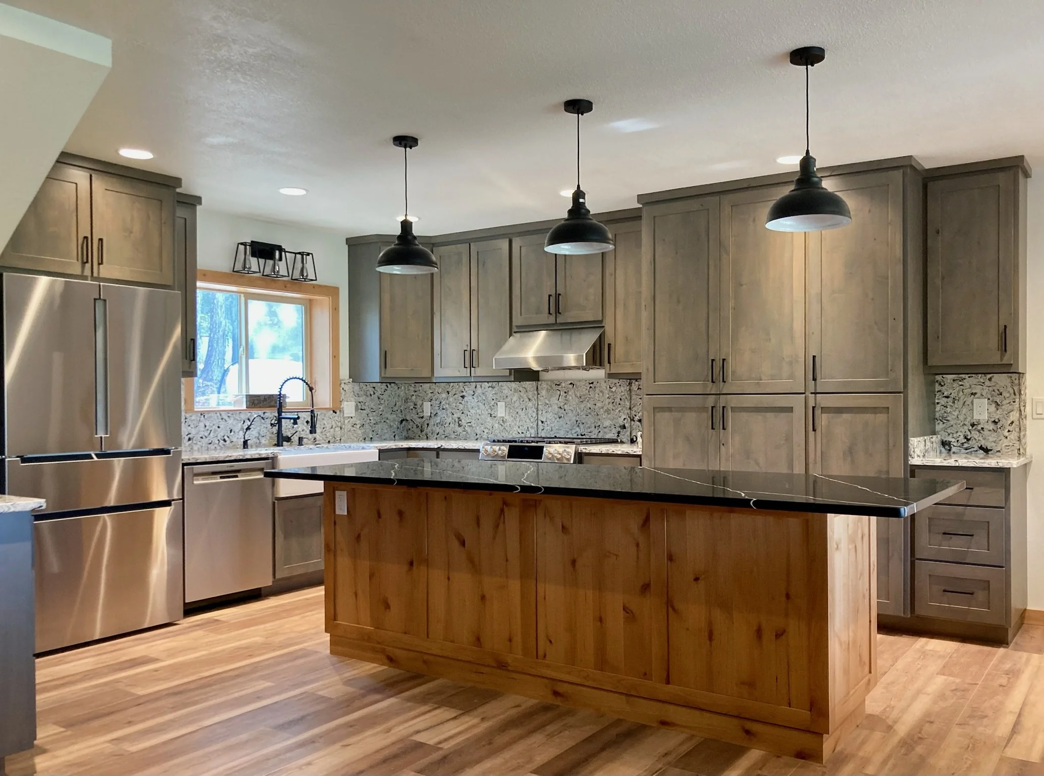 Modern kitchen with wooden cabinets, granite countertops, stainless steel appliances, island with wood finish, black pendant lights, and hardwood flooring.