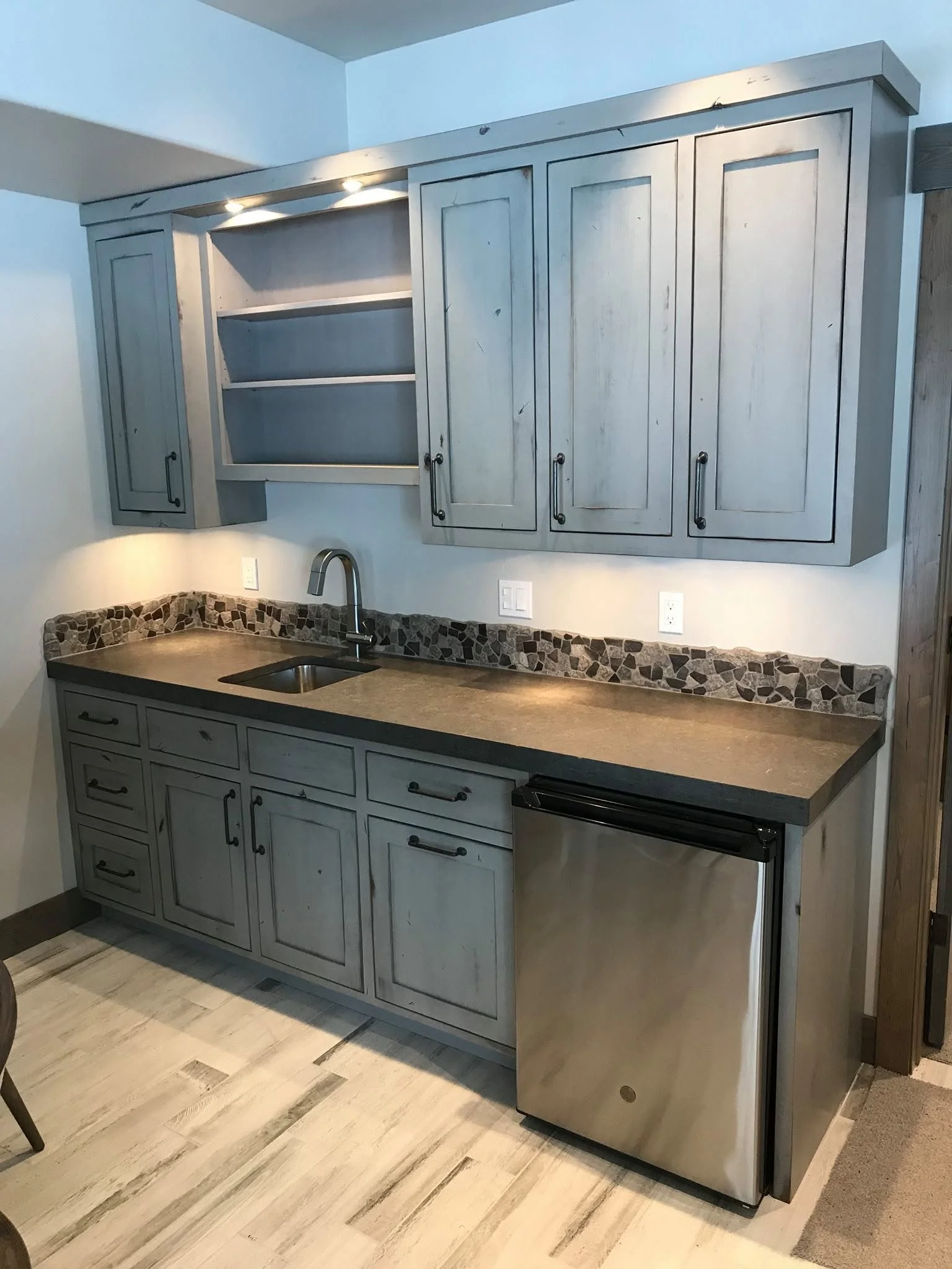 A kitchen with light grey cabinets above and below a countertop with a mosaic tile backsplash, a stainless steel sink with a curved faucet, and a small stainless steel refrigerator under the counter. There are electrical outlets on the wall, and part