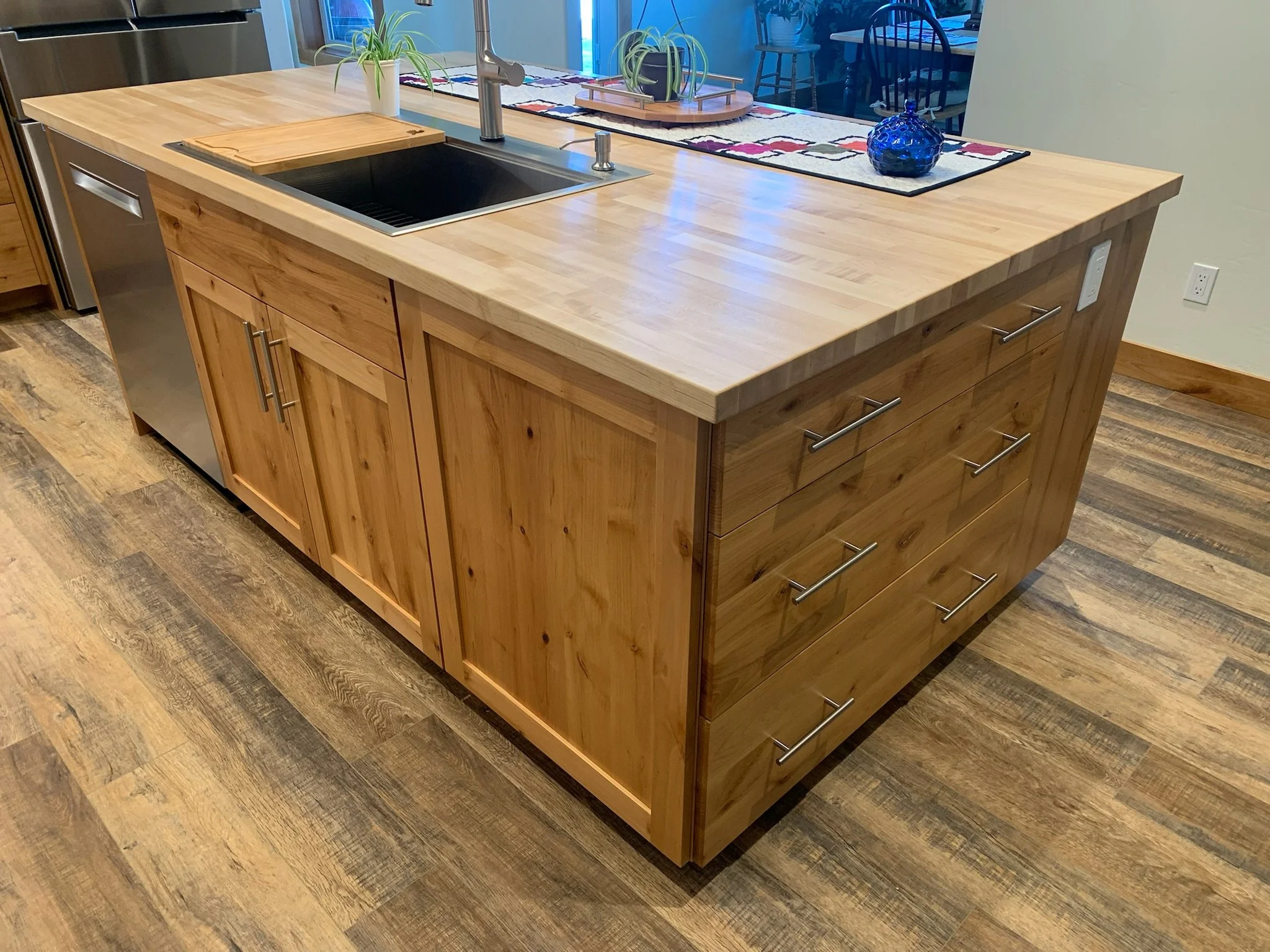 A kitchen island made of light-colored wood with multiple drawers and cabinets. There is a cutting board on top of the island, a kitchen sink with a faucet, and decorative items including a plant and a blue glass vase. The background shows a living o