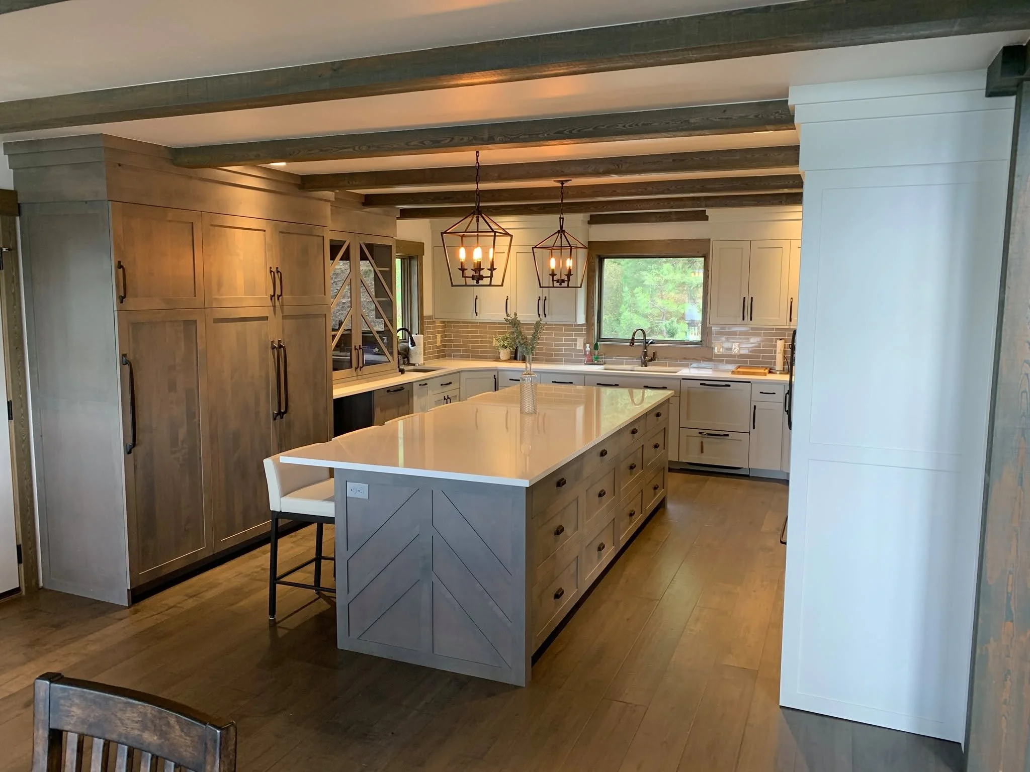 Modern kitchen with light wood cabinets, white countertop, brick backsplash, central island, two geometric pendant lights, window showing outside greenery, wooden flooring, and a wooden dining chair in the foreground.