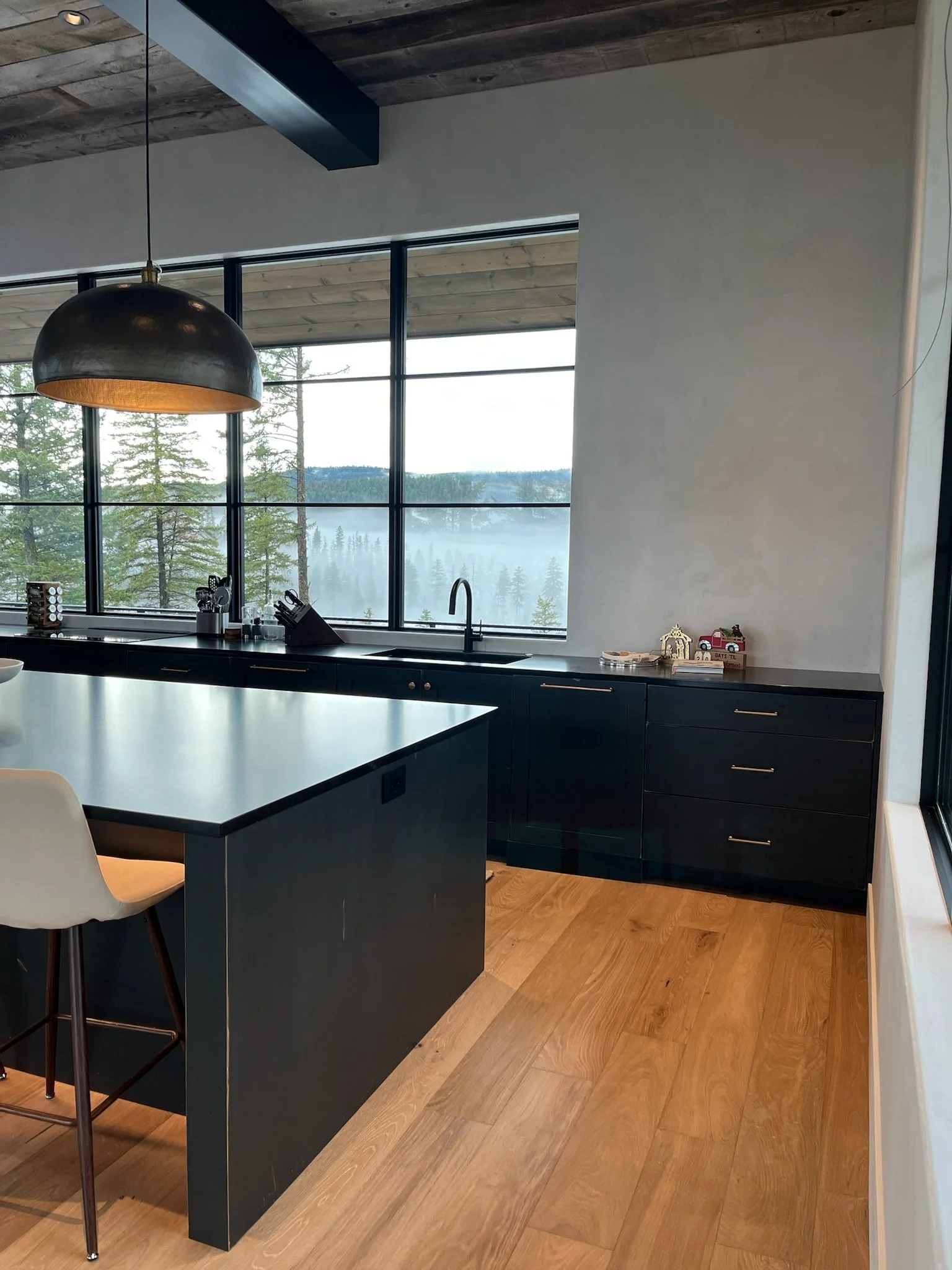 Modern kitchen with large window overlooking trees and lake, featuring black cabinets, a kitchen island, a hanging pendant light, and wooden flooring.