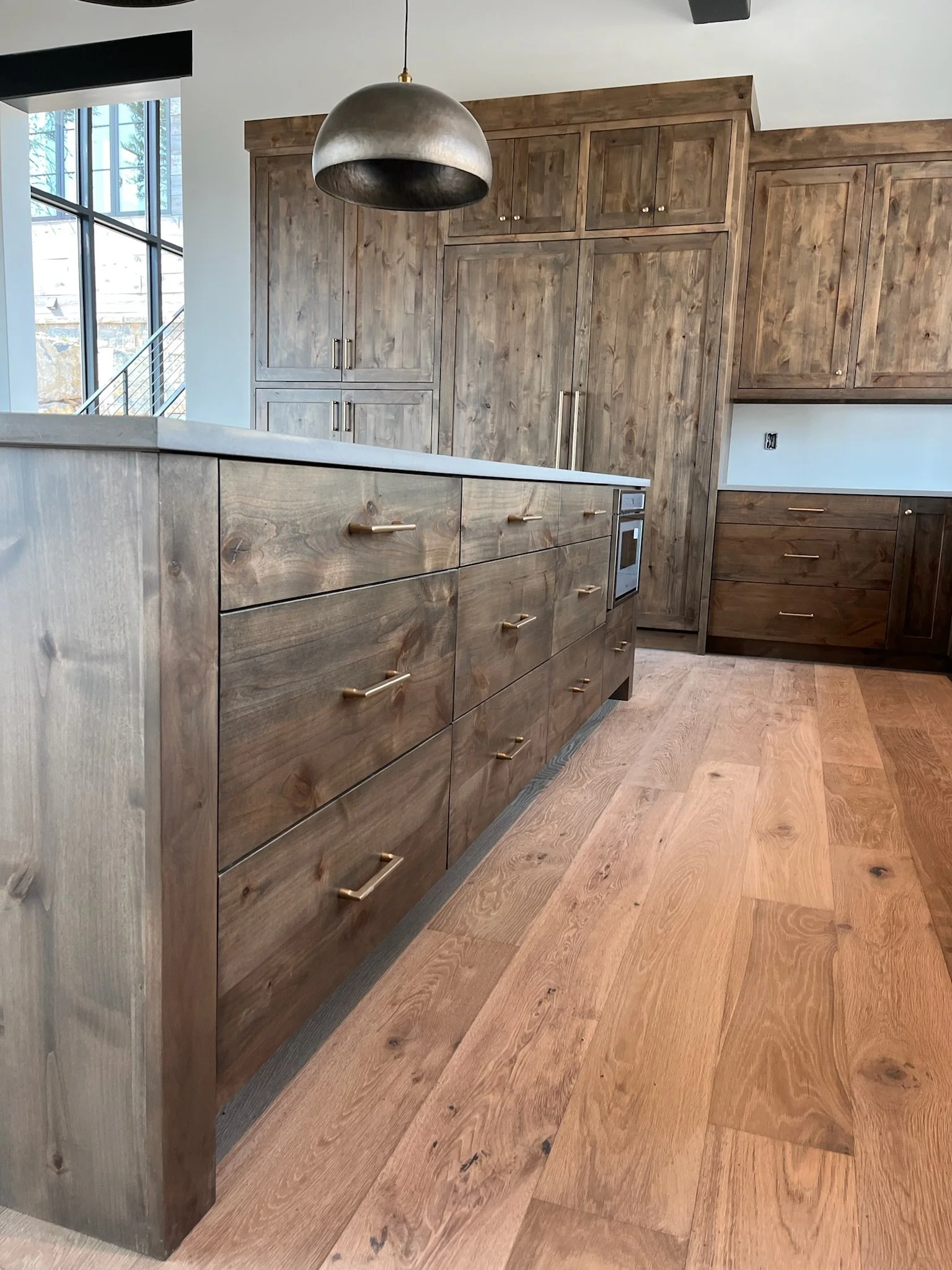 A kitchen with wooden cabinets, a kitchen island with drawers and gold handles, and a large window with natural light. The floor is also wooden.