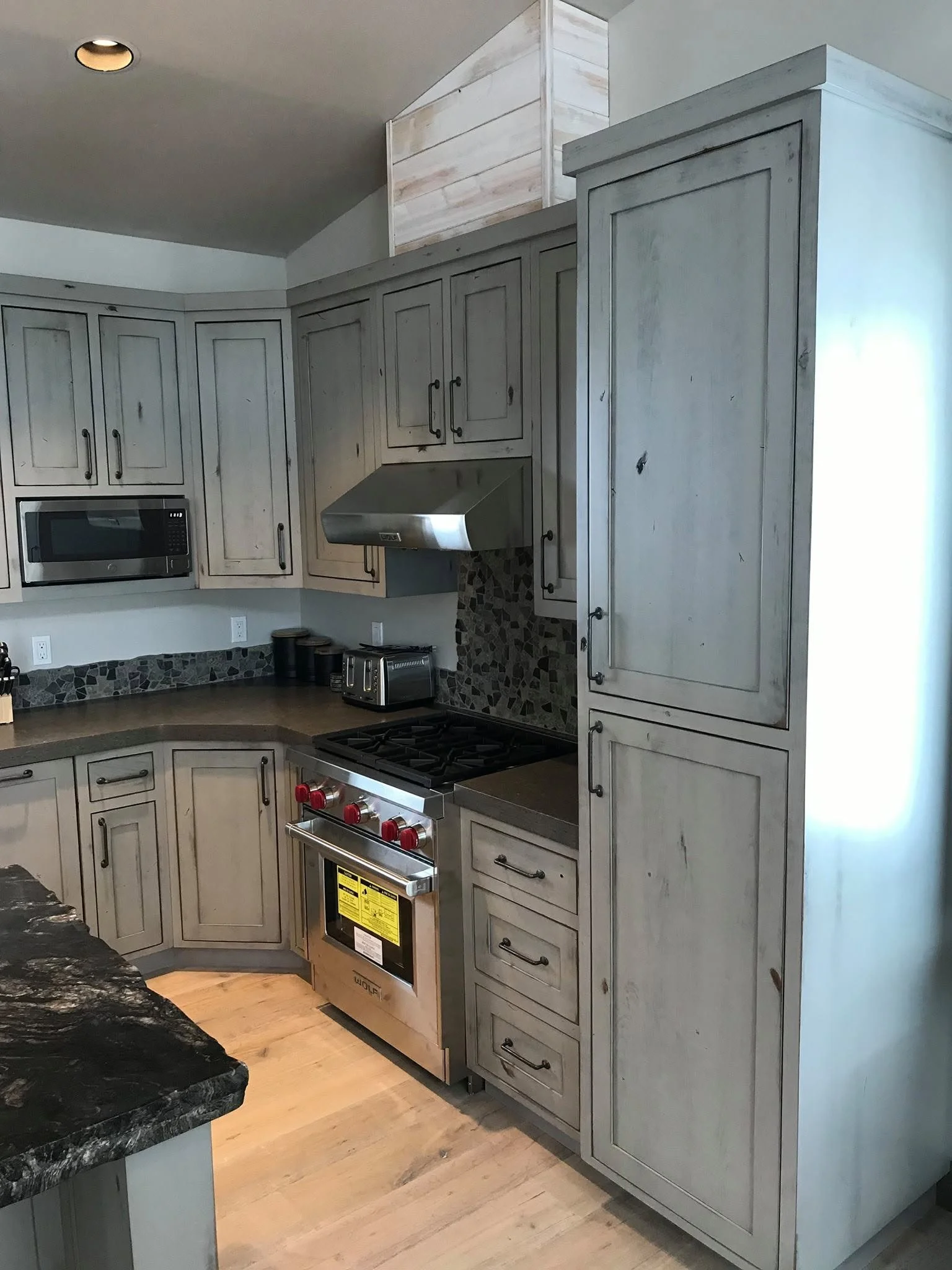 Kitchen with light gray wooden cabinets, stainless steel range hood, black stone countertop, and a stainless steel oven with red knobs. The backsplash has a mosaic tile pattern.