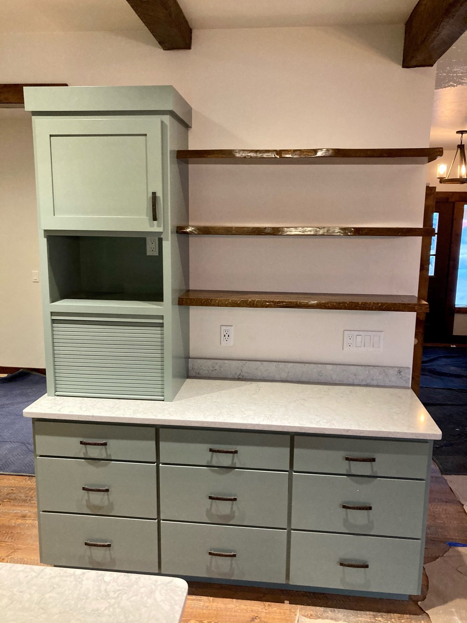 A kitchen cabinet with drawers and cupboards in a lime greenish color and brass handles, a marble countertop, and three wooden shelves mounted on a beige wall.