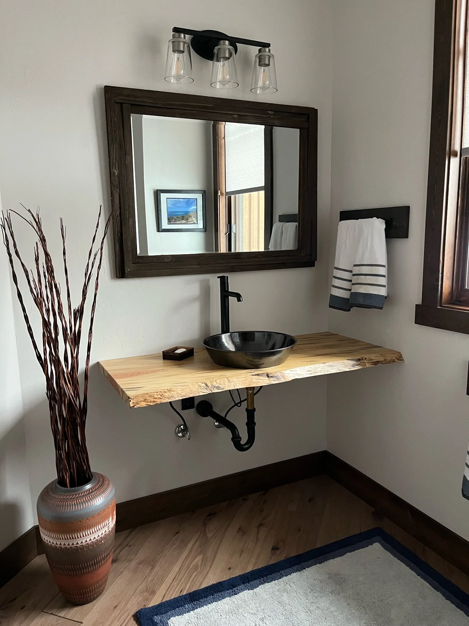 Bathroom vanity with a natural wood countertop, black vessel sink, black faucet, rectangular flush mount mirror with a dark wood frame, wall light fixture with three clear glass shades, and a ceramic decorative vase with tall dry branches.