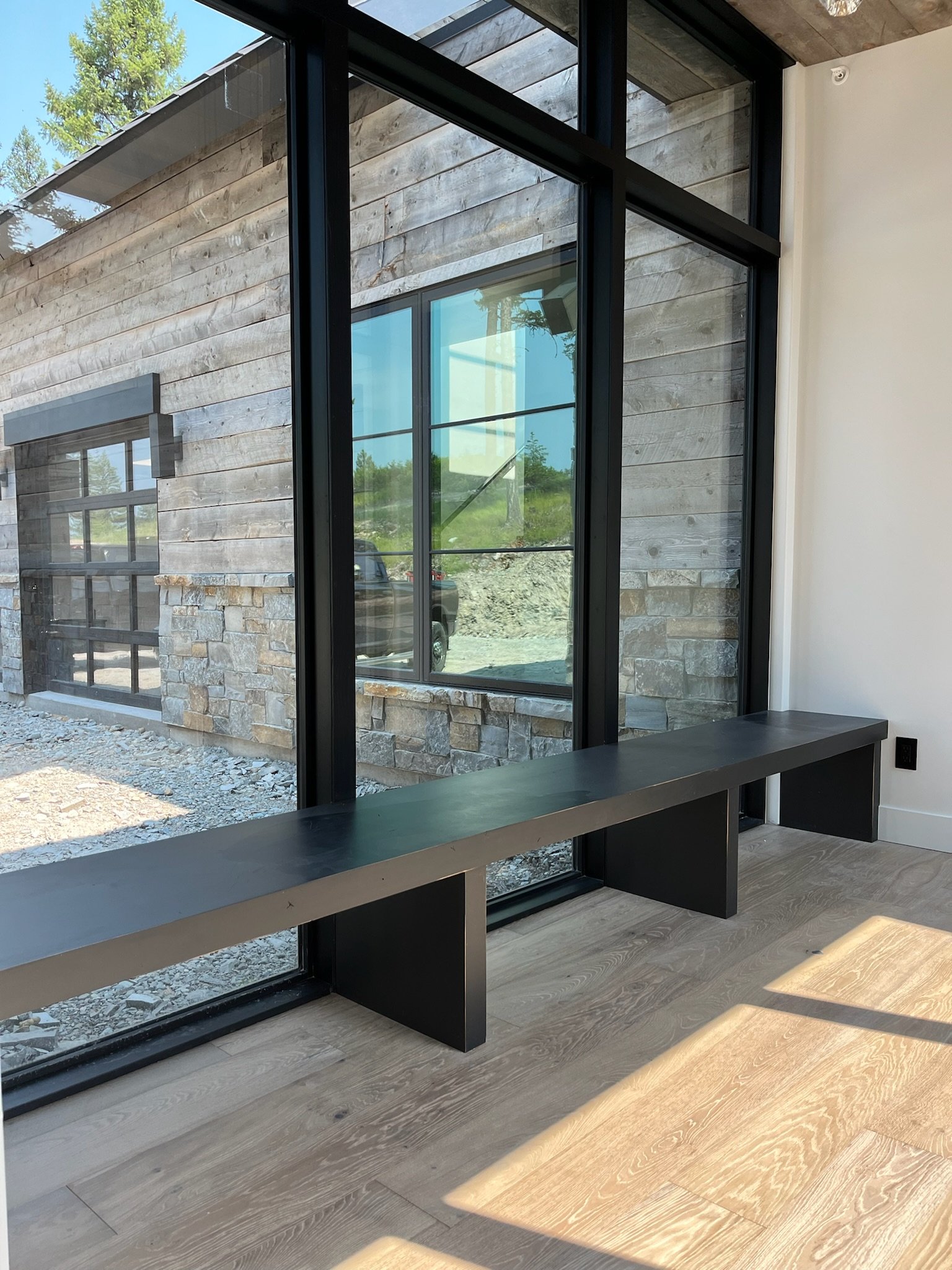 A modern interior space with large floor-to-ceiling windows, a black window frame with multiple panes, wooden flooring, and a view of the outdoors with trees and a parked vehicle.