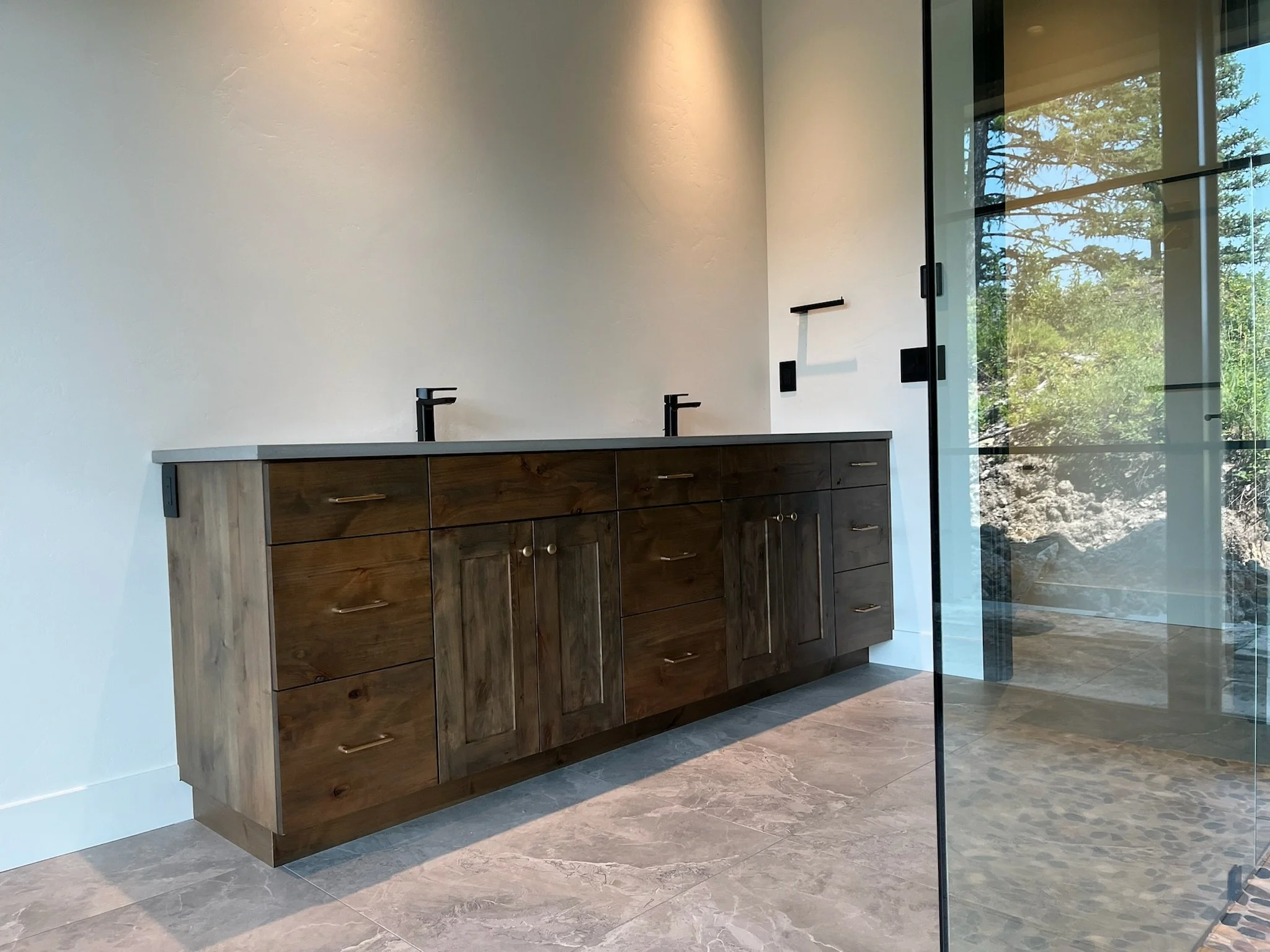 Modern bathroom with a light-colored wall, dark wood vanity with multiple drawers, black faucets, and a large glass window showing trees outside.