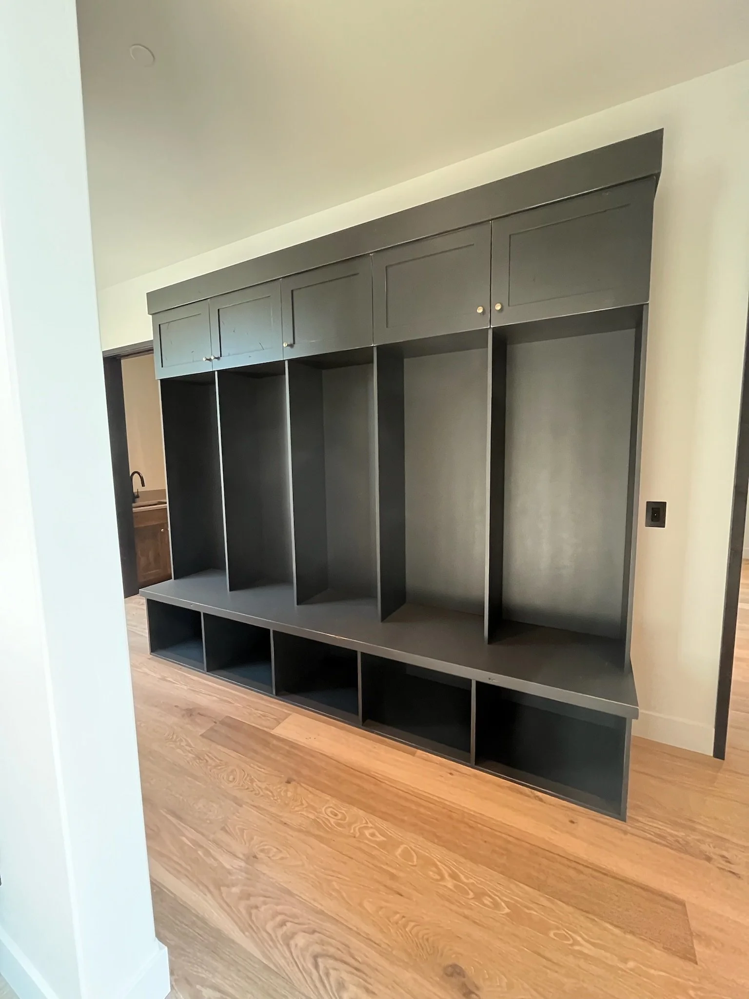 A black wall-mounted storage unit with closed cabinets on top a large open shelving area with cubbies, set against a white wall in a room with wooden flooring.