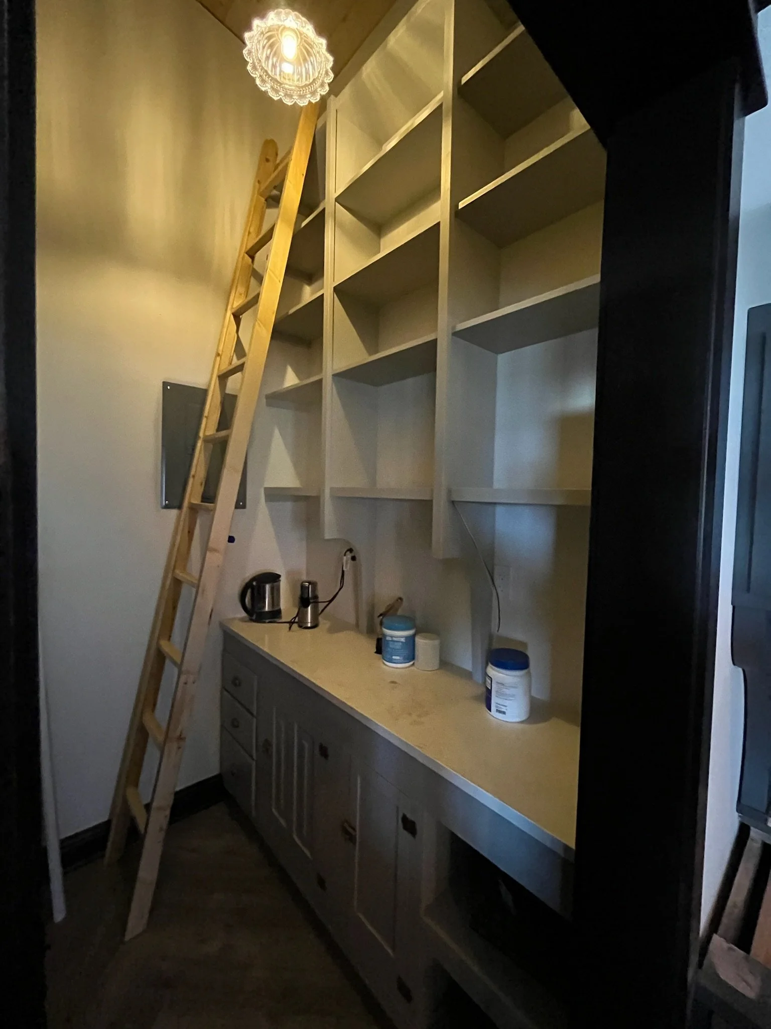 A small kitchen or utility area with a gray cabinet with cupboards and a beige countertop. Above the counter, there are open shelves, and a wooden ladder leans against the wall. On the countertop, there is a kettle, a blender, and containers of powde