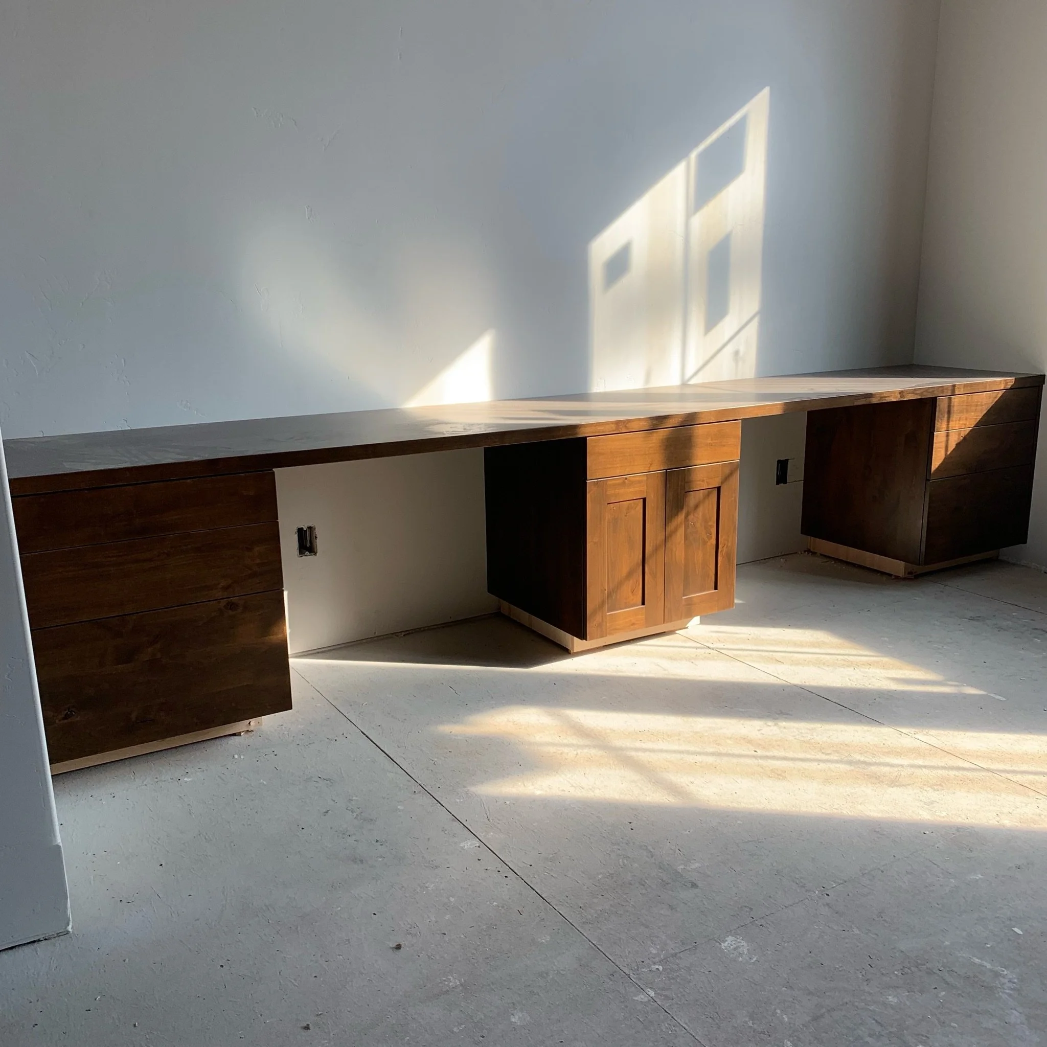 A wooden countertop or table with a light-colored, smooth surface, placed against a plain, light-colored wall. Sunlight is casting shadow patterns on the wall and the countertop.