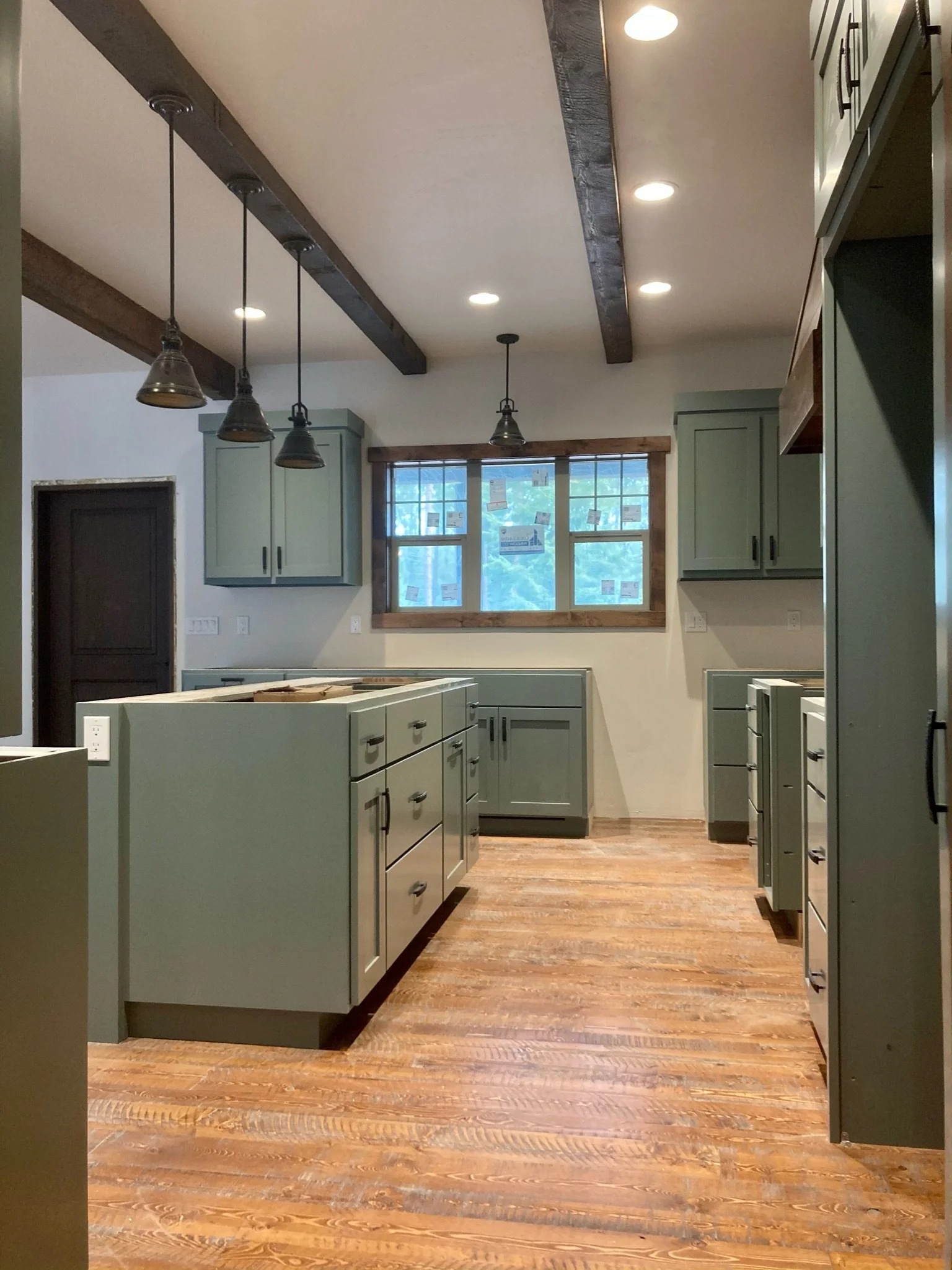 This image shows a kitchen with light green cabinets, a large window with wooden trim, a wood floor, and exposed ceiling beams. There are three pendant lights hanging from the ceiling.