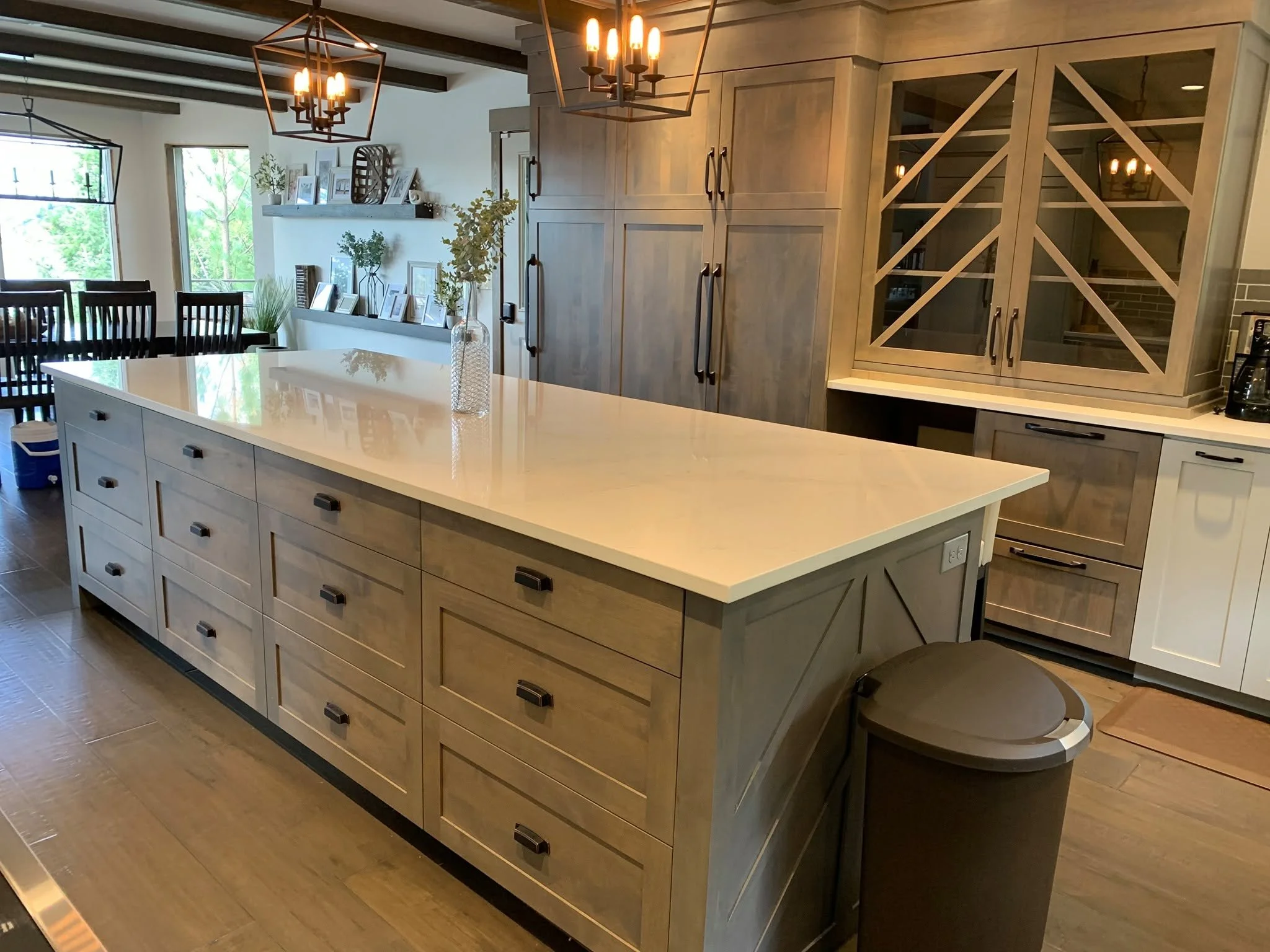 A modern kitchen with a large white island countertop, wood cabinetry, black handles, and a built-in wine rack, illuminated by geometric pendant lights.