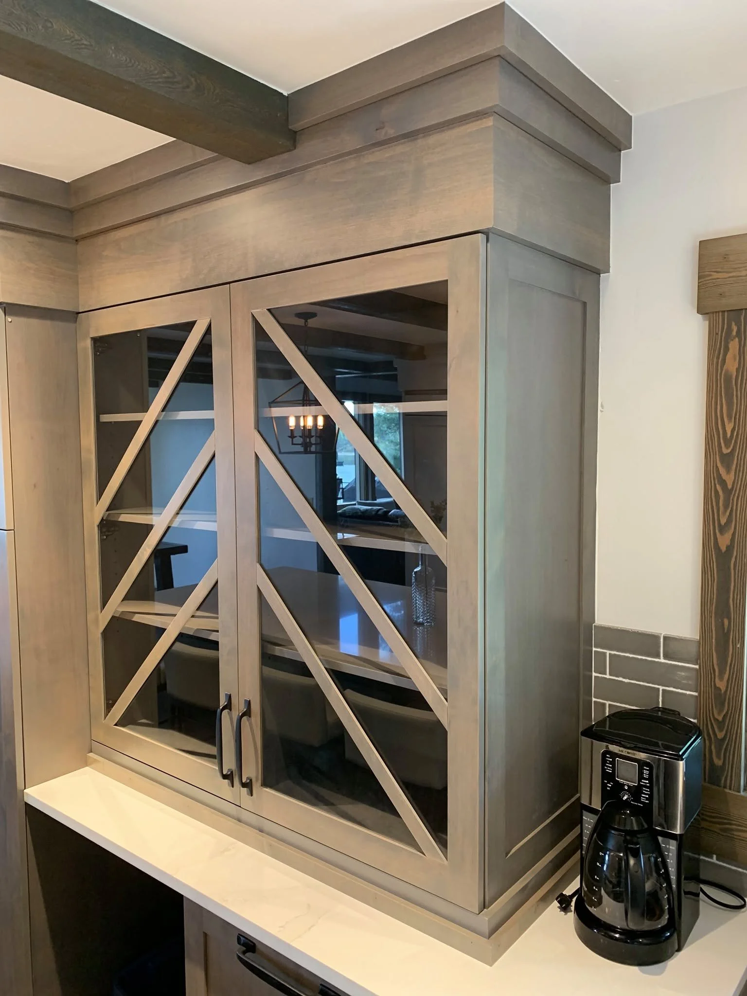 A wooden kitchen cabinet with glass doors and a crisscross design, placed on a white countertop. A coffee maker with a glass carafe is positioned nearby, and a black container is on the countertop. The cabinet is against a wall with a wooden trim and