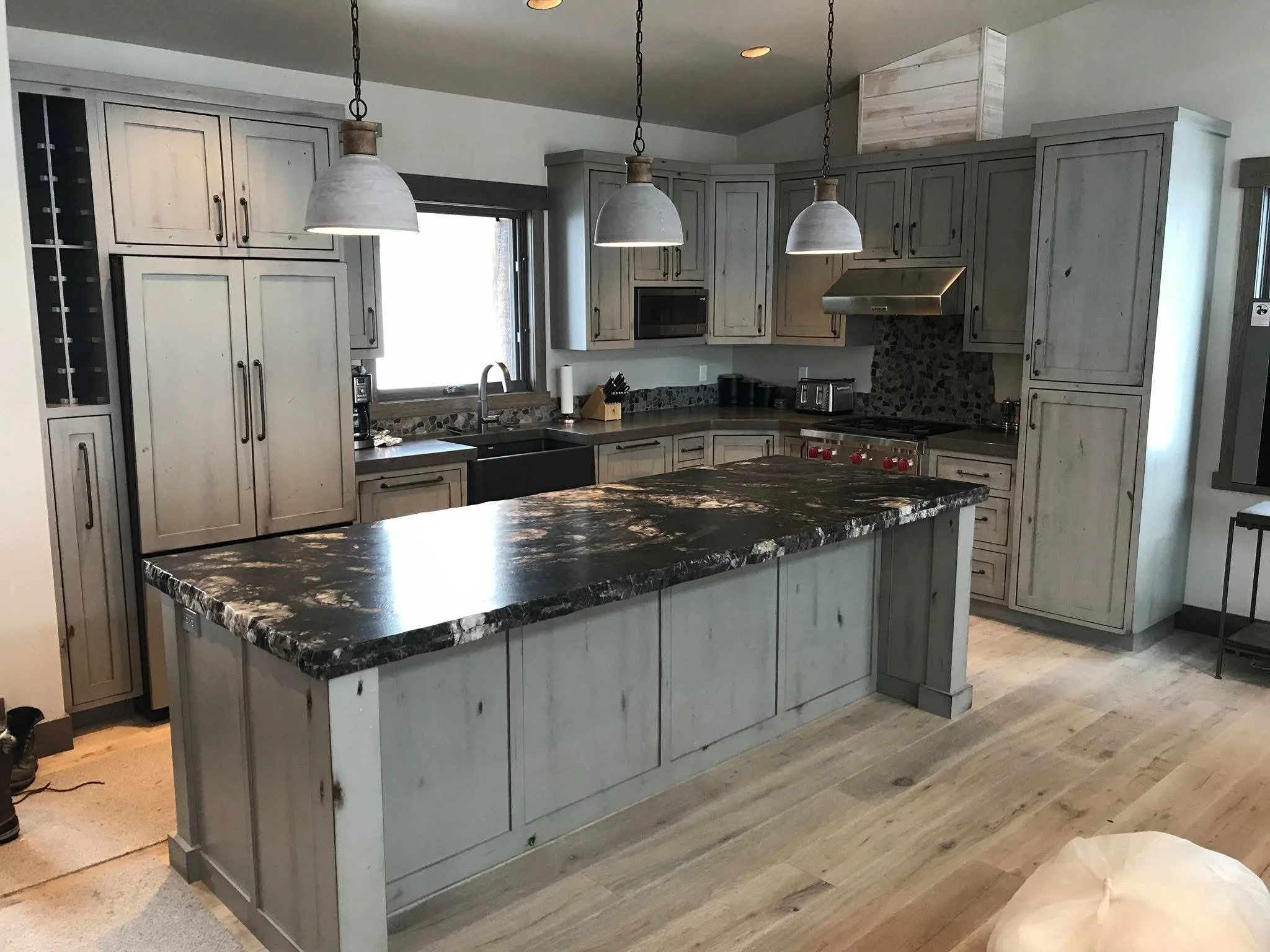 A spacious kitchen with white wooden cabinets, a dark granite island, stainless steel appliances, and pendant lights hanging over the island. There is a window above the sink and various kitchen essentials on the countertop.