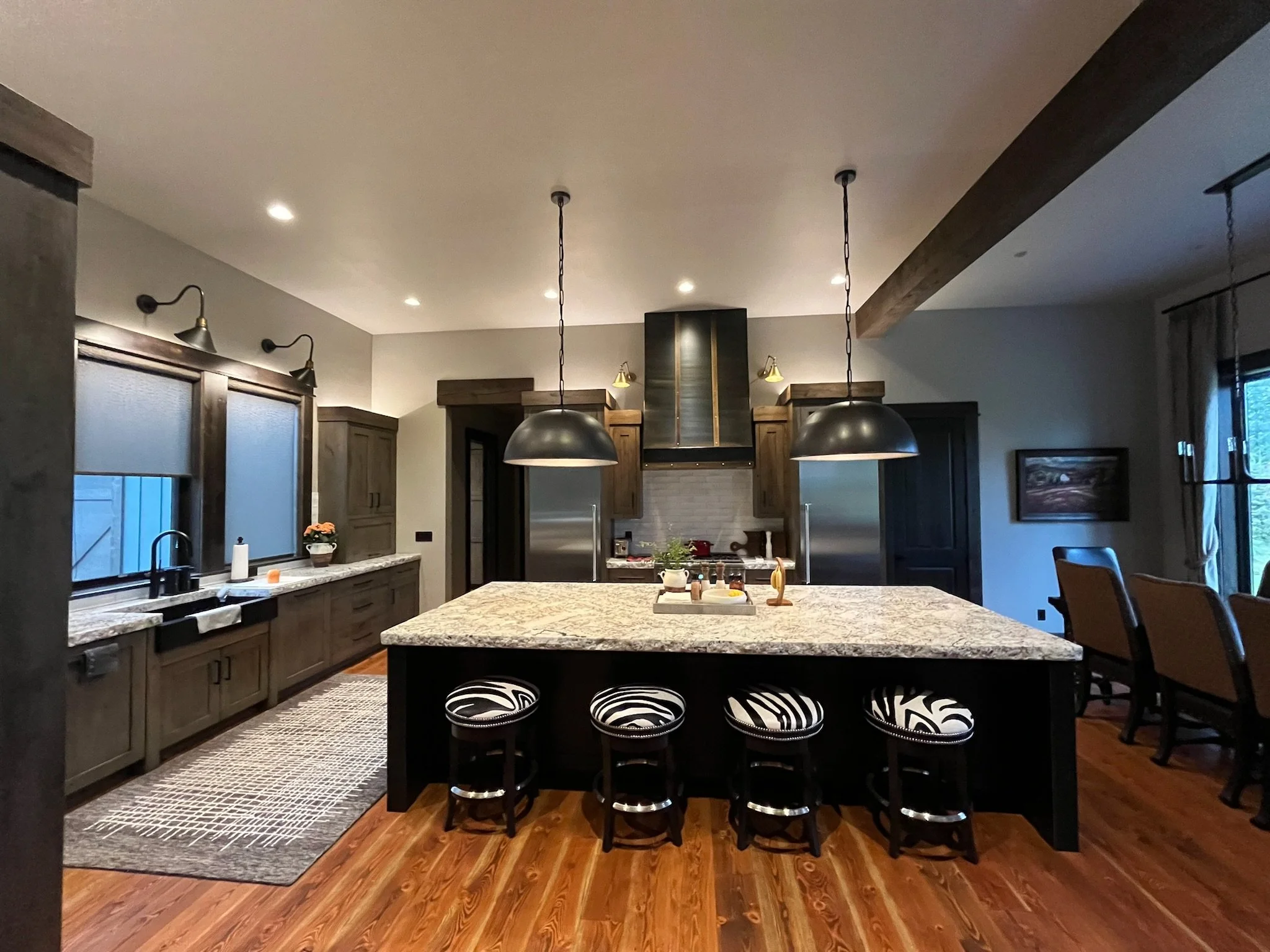 A spacious kitchen with dark wooden cabinets, a large granite island, and black-and-white zebra print bar stools. Pendant lights hang over the island, and there are windows with shades on the left. The floor is wood, and there is a gray patterned rug