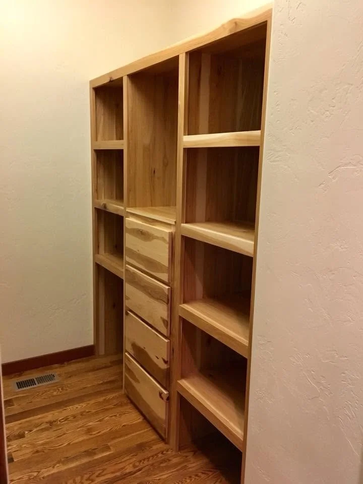 Unfinished wooden shelving unit with multiple open shelves and four drawers, located in a corner of a room with hardwood flooring and beige walls.