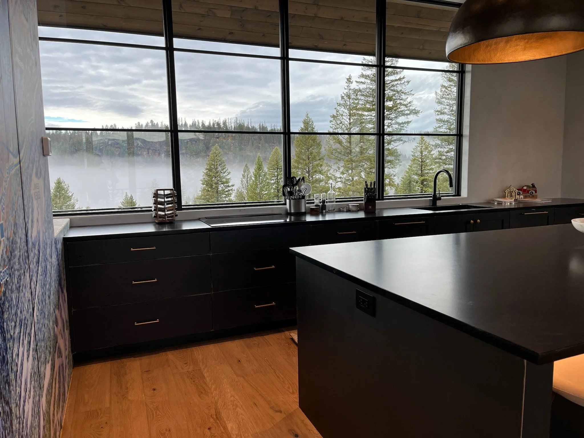 Interior view of a modern kitchen with large windows showing a foggy forested landscape outside. The kitchen has black cabinets, a black countertop, and various kitchen utensils and containers on the counter.