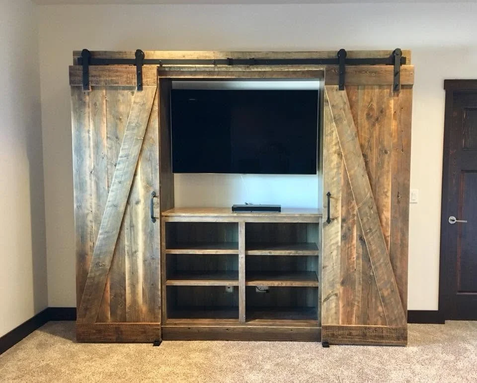 A rustic wooden barn door TV cabinet with sliding doors on a black track, surrounding a flat-screen TV mounted on a wall, with open shelves below and a small cable box on top.