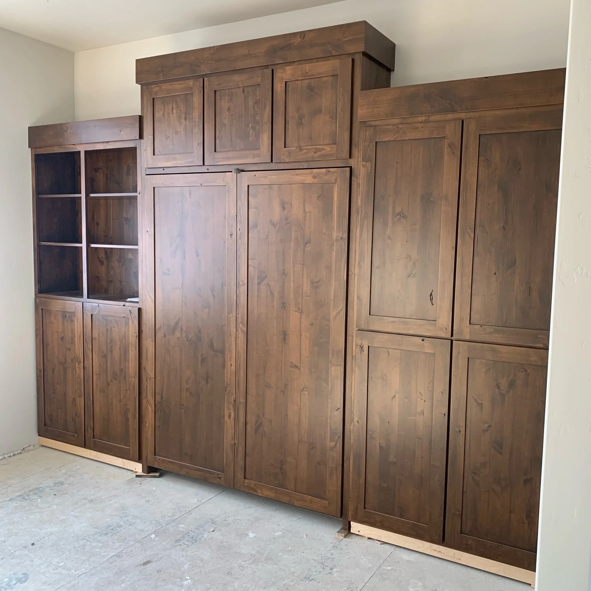 Unfinished wooden wardrobe or cabinet with multiple compartments and doors, standing on a concrete floor in a room under construction.