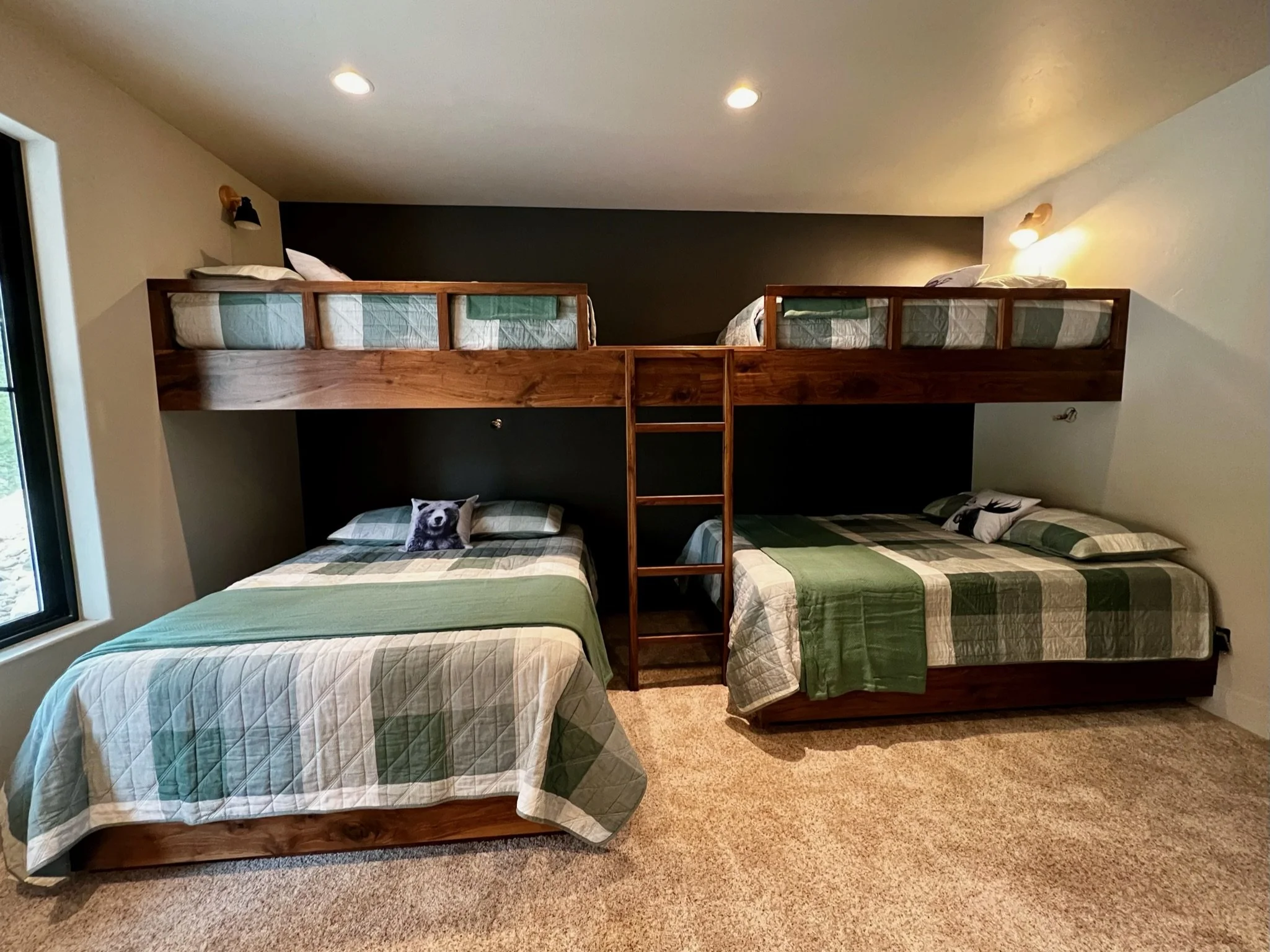 A bedroom with two queen beds, one with a dog pillow, and a bunk bed with a ladder, all covered with green and beige plaid quilts. There's a small window on the left and ceiling lights, with wall-mounted lights above each bed.