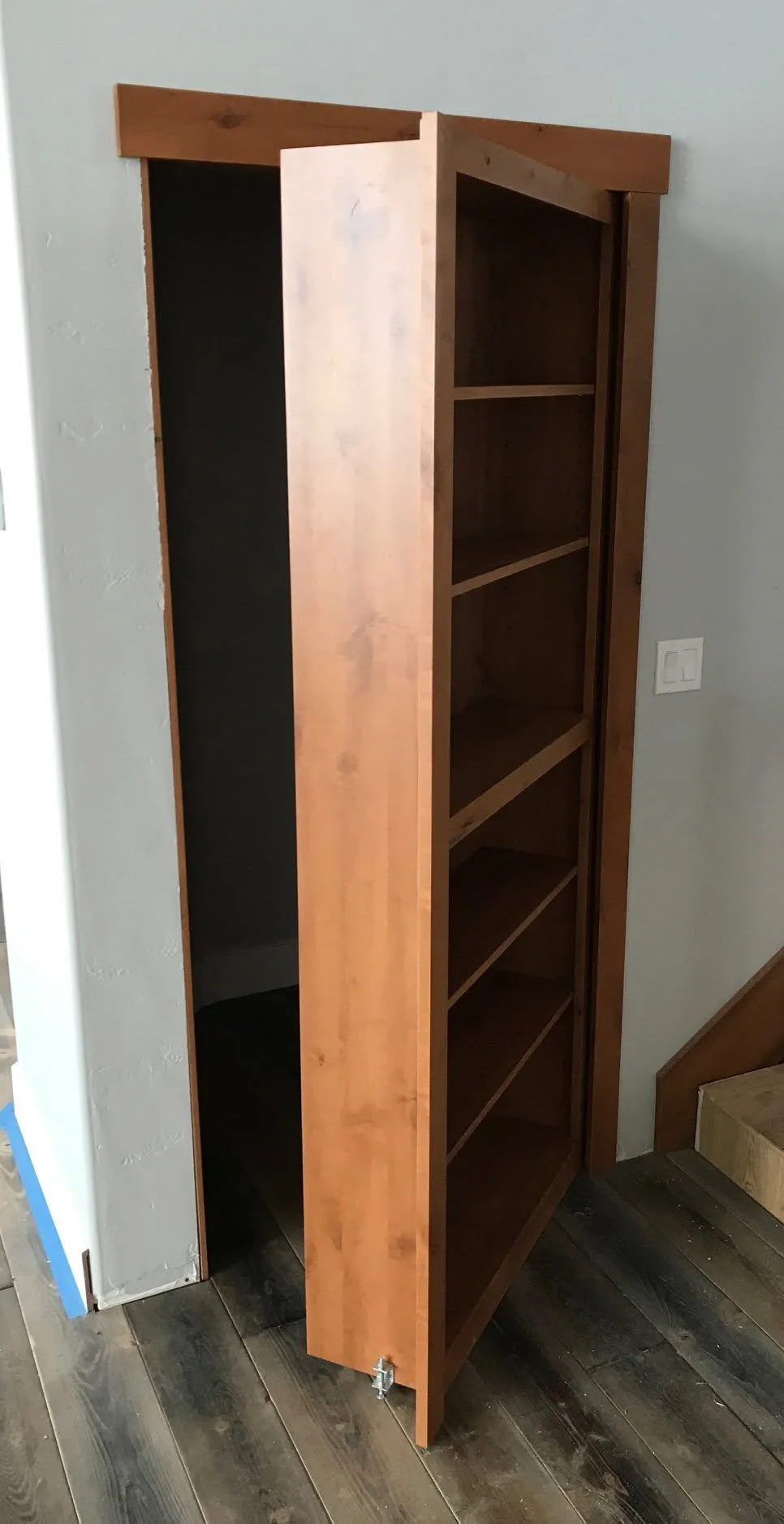 A wooden bookshelf with four empty shelves, standing next to a light gray wall and near a staircase with wooden steps.